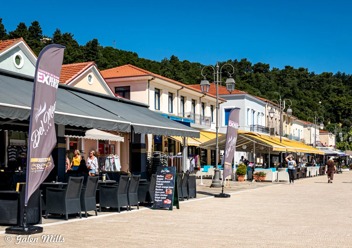 Outdoor cafe and shops on a street with people walking, trees in the background, and colorful buildings with red and yellow awnings.