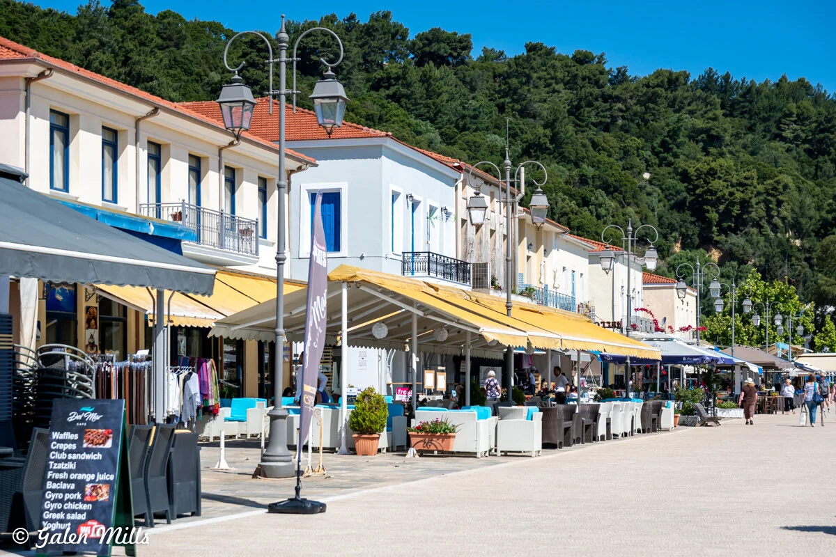 Outdoor cafes with yellow awnings, shops, and people walking on a sunny day, lined by trees.