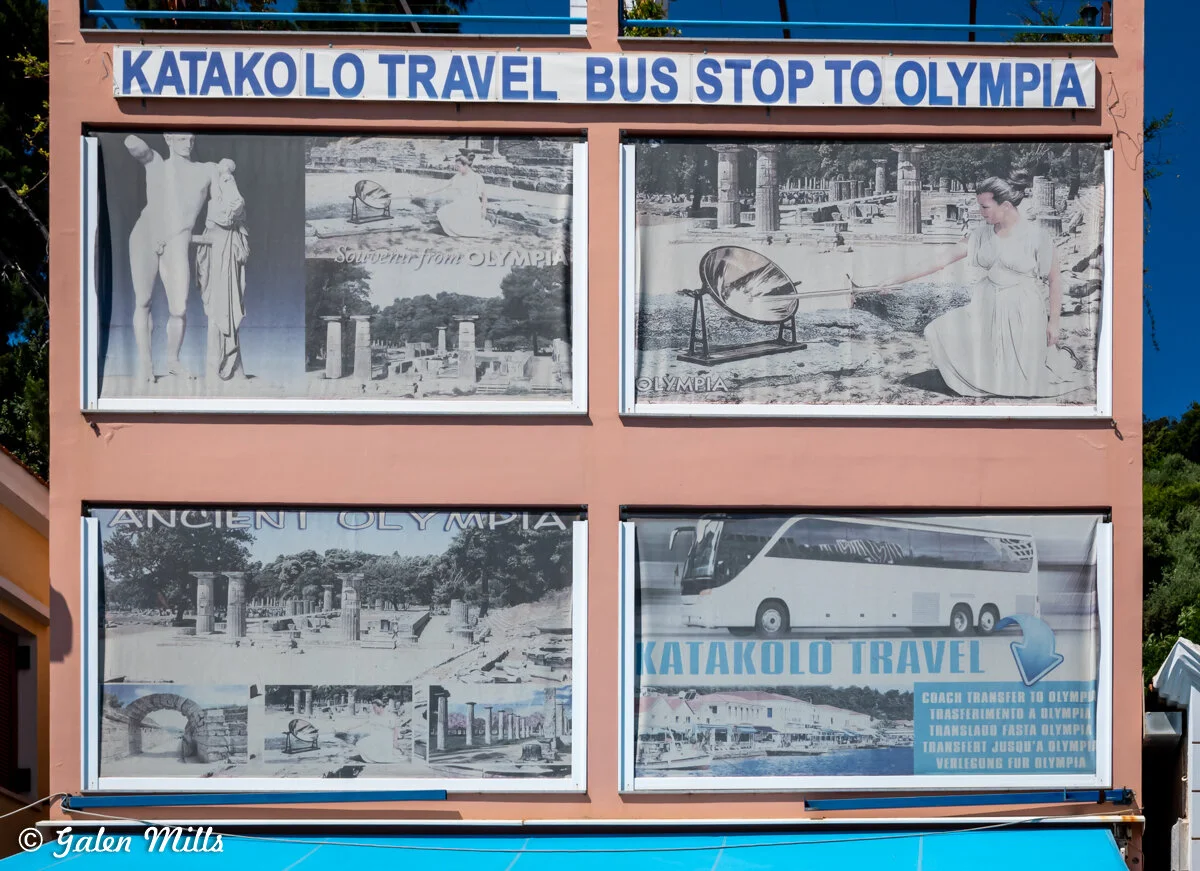 Travel agency sign for Katakolo Travel, advertising bus tours to Olympia, featuring images of ancient Olympia sites and a bus.