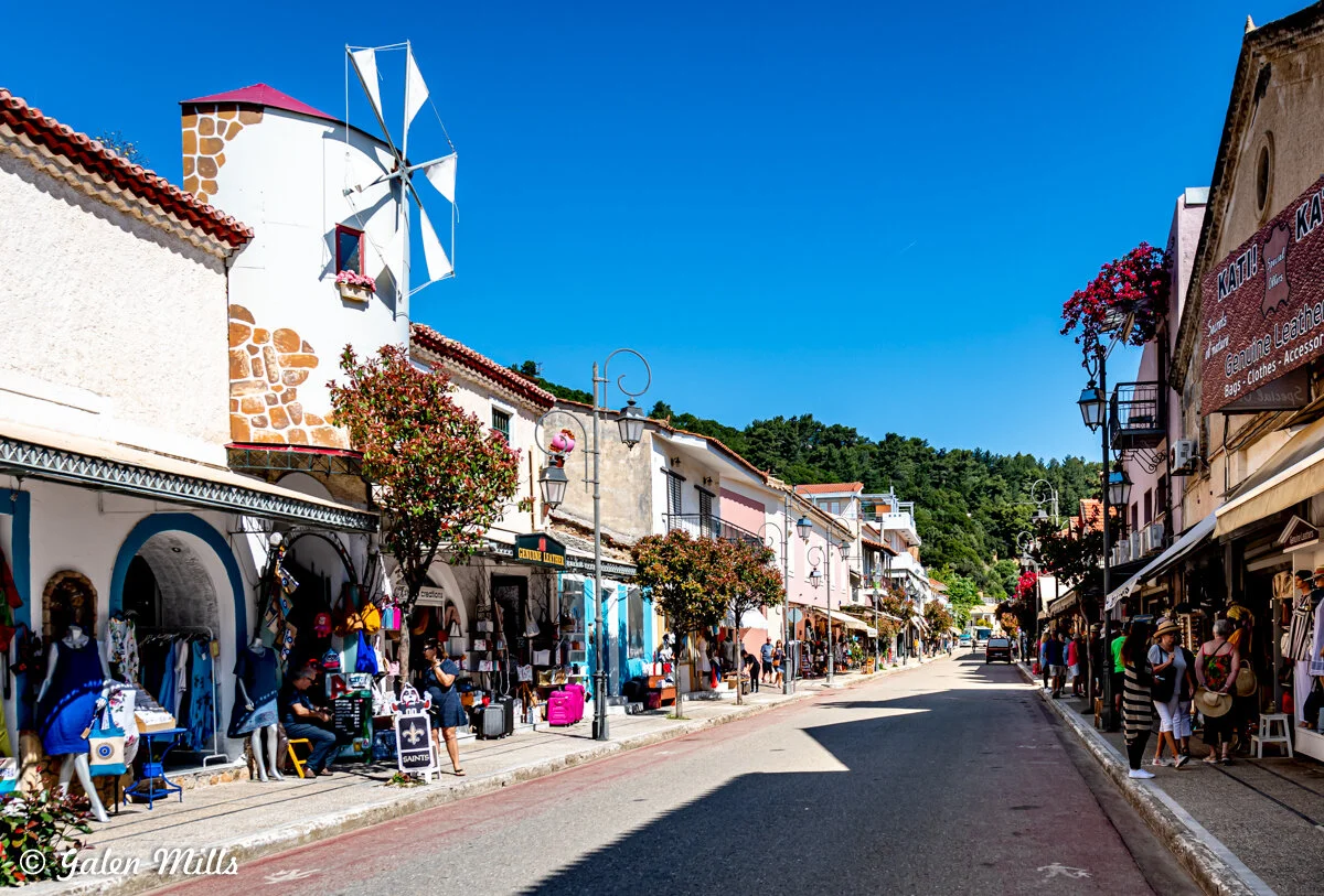 Main street in a Greek town with traditional buildings, shops, a windmill, and pedestrians.