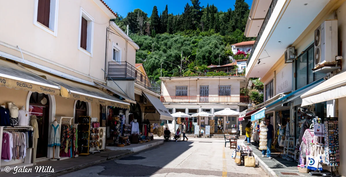 Street view of a small shopping area with clothing stores, boutiques, and outdoor seating. The shops have awnings, and there are trees and hills in the background.