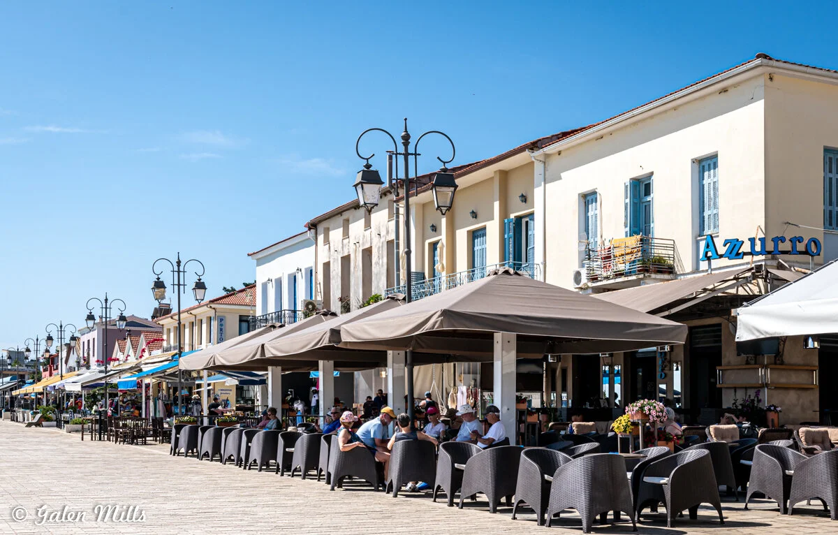 Coastal promenade with outdoor cafes and colorful buildings, featuring wicker chairs and umbrellas, on a sunny day.