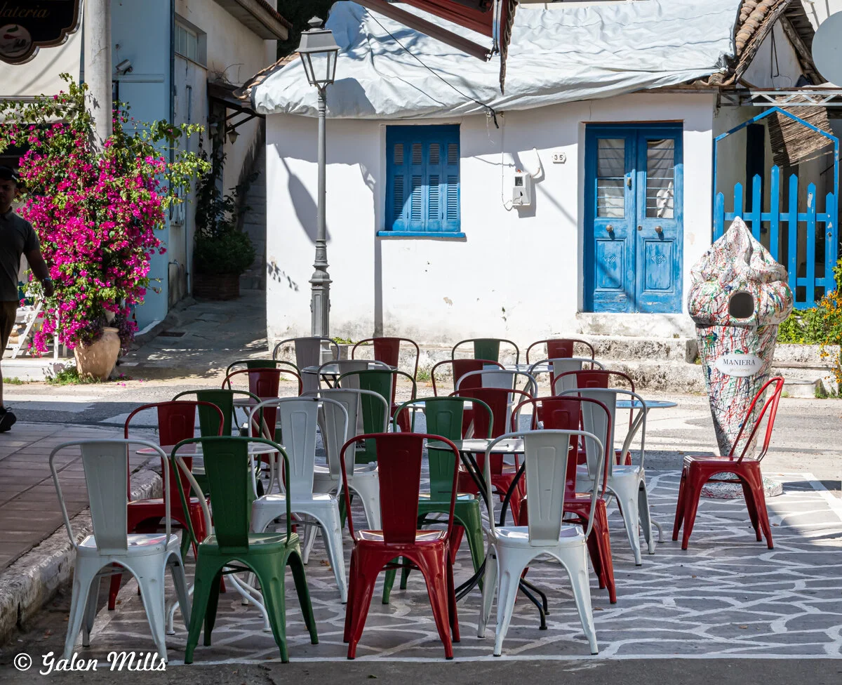 Outdoor seating area in a village with metal chairs in red, white, and green, a large ice cream cone statue, pink bougainvillea, white buildings with blue doors and windows, and a lamppost.