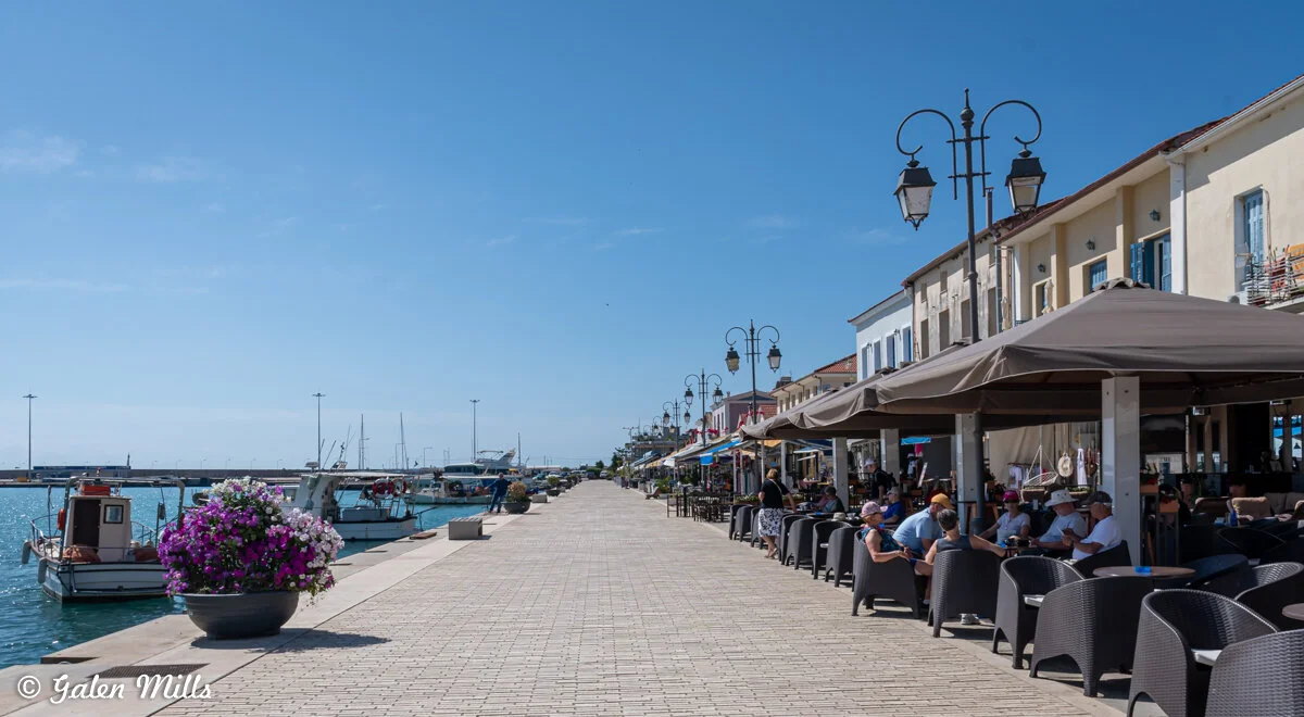 Seaside promenade with outdoor cafes and moored boats on a sunny day.