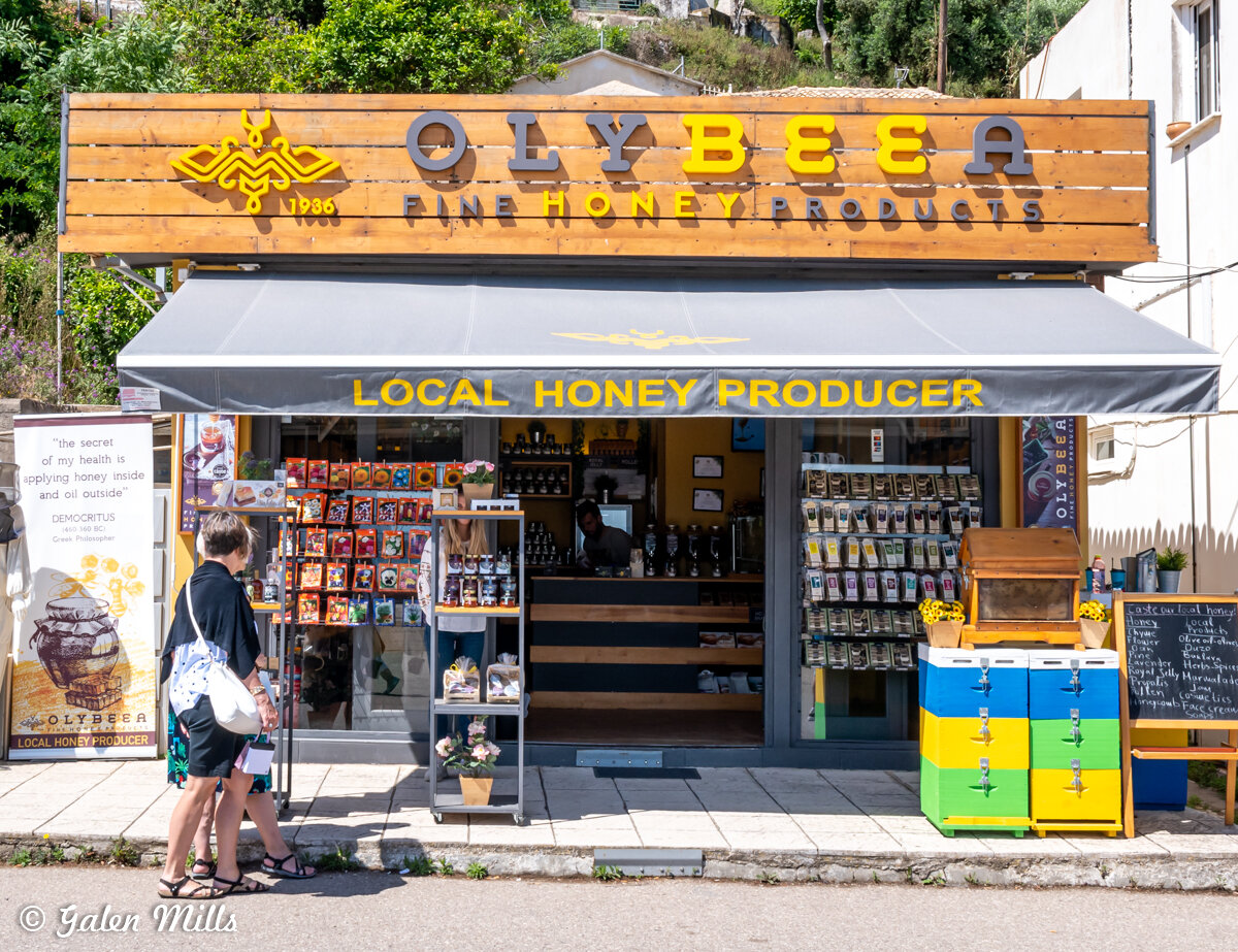 Front view of Olybeea honey shop with wooden sign and outdoor display of honey products, featuring a gray awning labeled 'Local Honey Producer' and two people browsing.