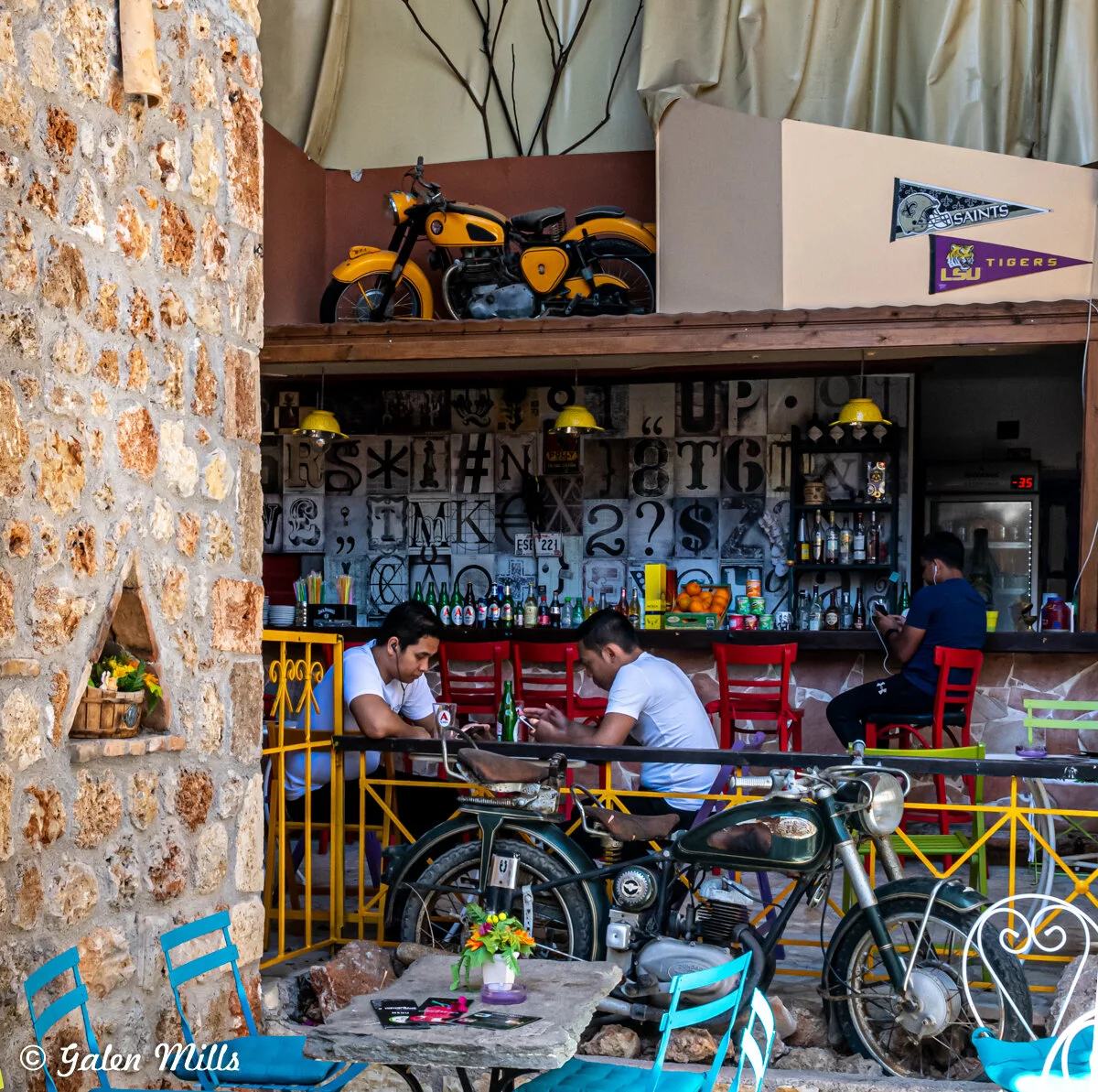 Outdoor café scene with people sitting at tables, a vintage motorcycle, colorful chairs, and a bar with bottles and decorations on the wall. A yellow motorcycle is displayed above the bar.