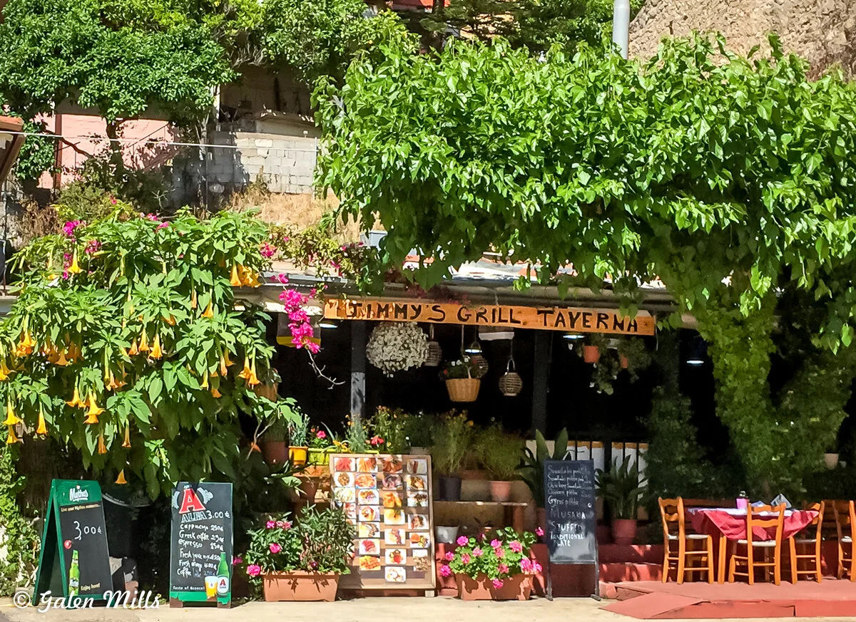 Outdoor view of a grill taverna with plants, handwritten menu boards, and tables with red tablecloths.