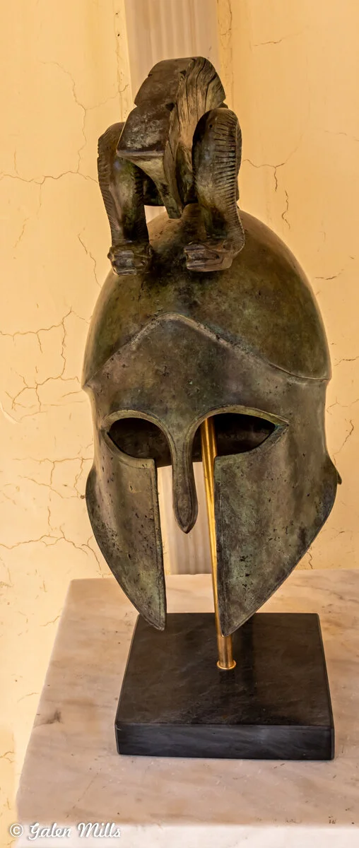Ancient Greek bronze helmet with a decorative animal figure on top, displayed on a pedestal.