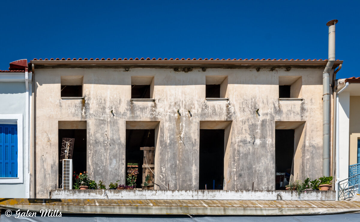 Old building with weathered facade, multiple square openings, a rooftop with red tiles, and potted plants on the ledge, against a clear blue sky.