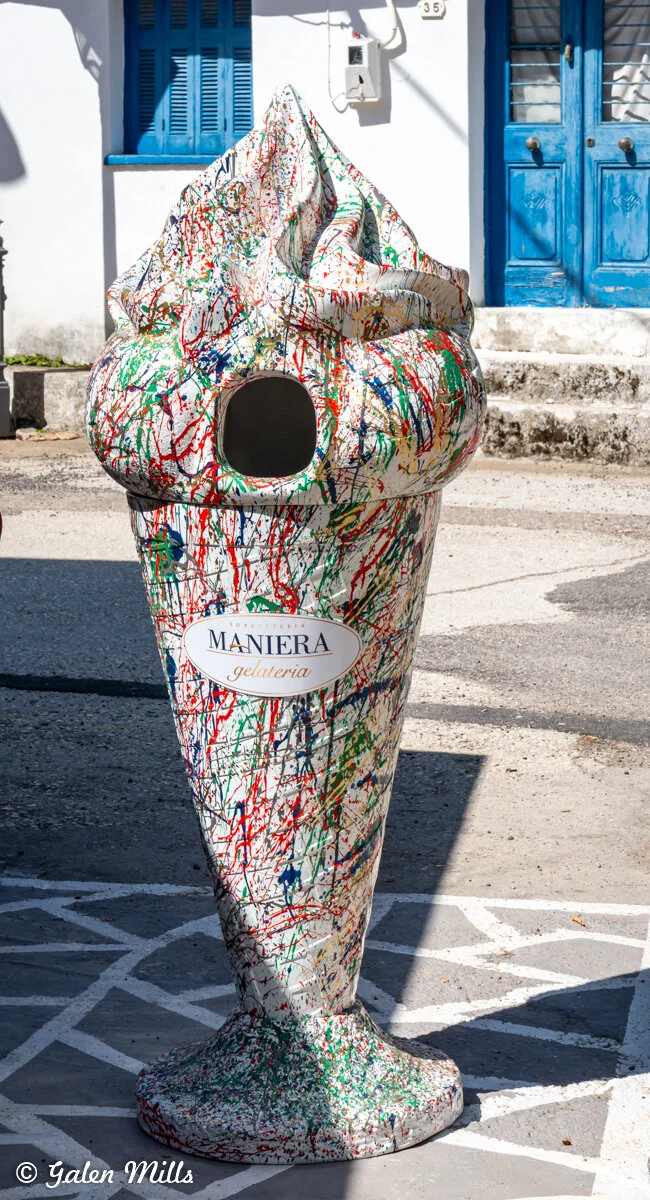 Colorful splatter-painted ice cream cone sculpture with a cutout, labeled 'Maniera Gelateria,' placed on a street with blue doors and windows in the background.