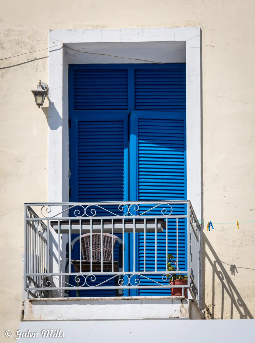 A small balcony with a metal railing and a wicker chair in front of blue shutters and a light-colored wall. A potted plant sits on the balcony. Clothespins hang on a line.