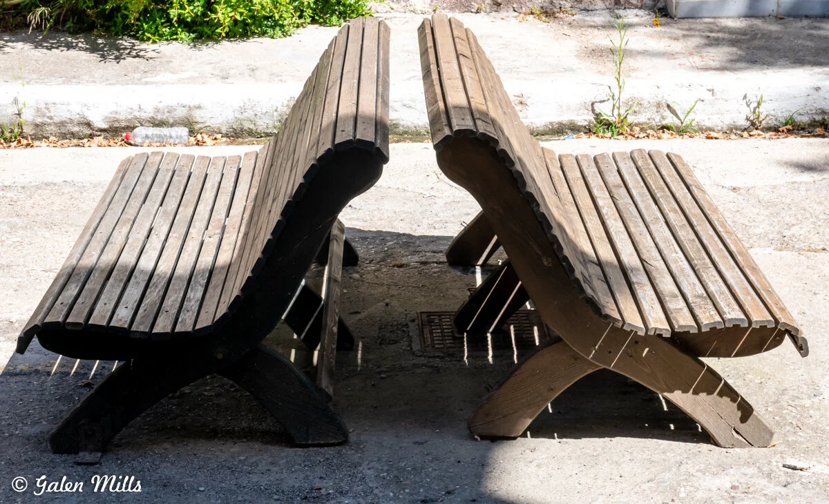 Two wooden curved benches placed back-to-back on a sidewalk, with sunlight casting shadows.