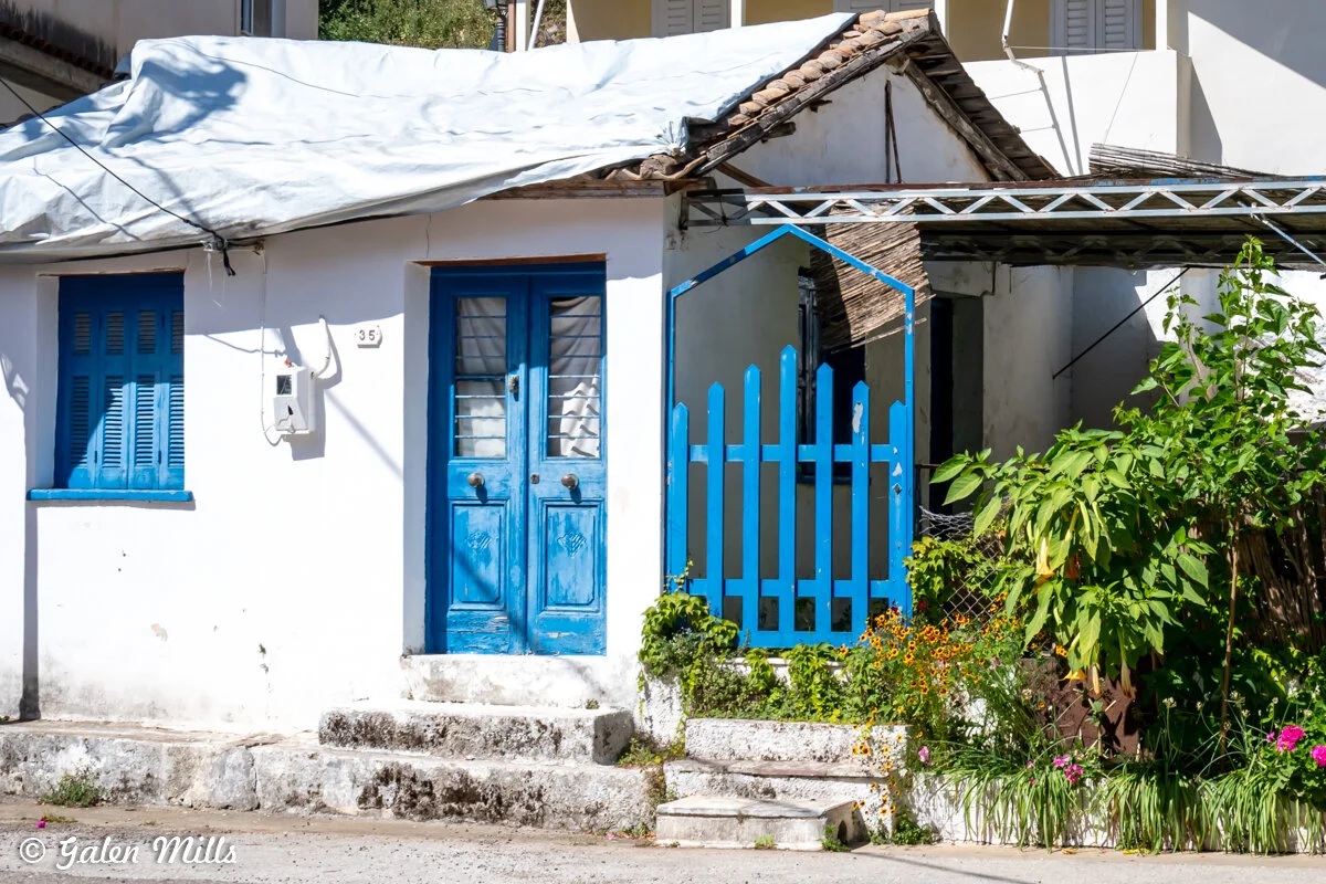 Small white house with blue doors and windows, a blue fence, and plants in the garden.