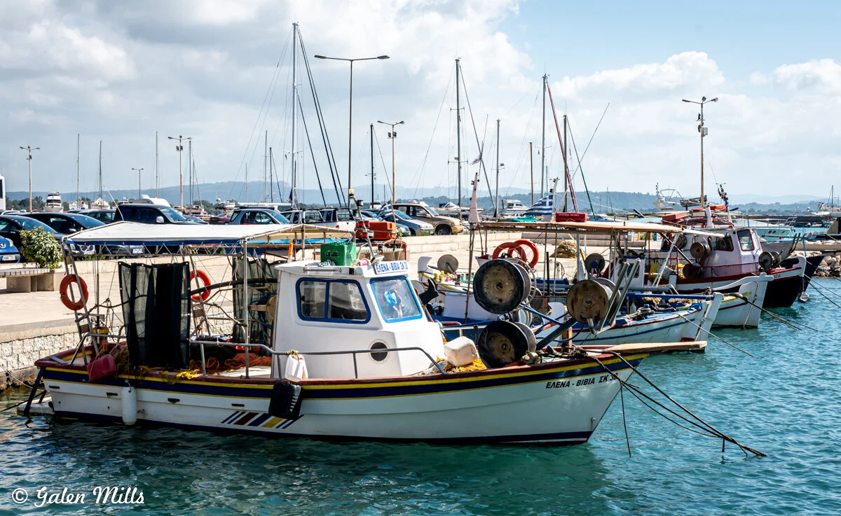 Fishing boats docked at a harbor, with parked cars and buildings in the background.