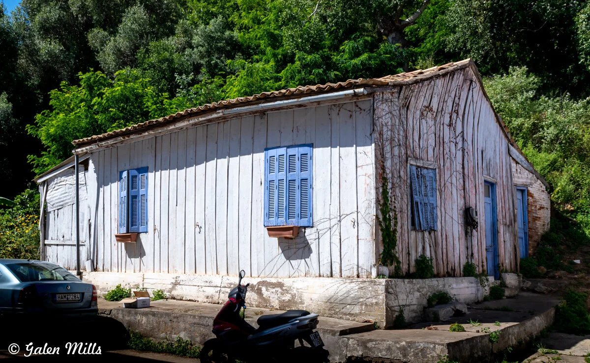 Rustic wooden house with blue shutters and doors, surrounded by greenery, a parked car, and a motorbike in front.