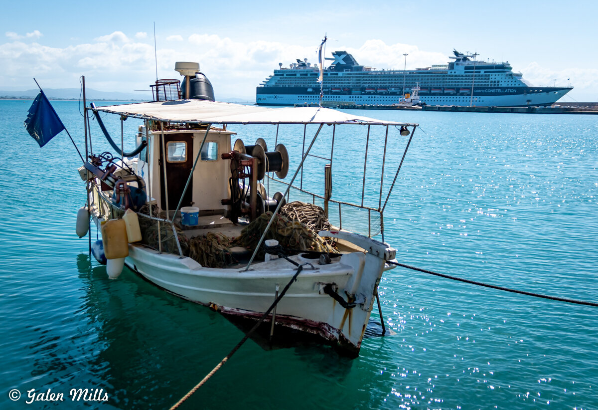 A small fishing boat docked in a harbor with a large cruise ship in the background on a sunny day.