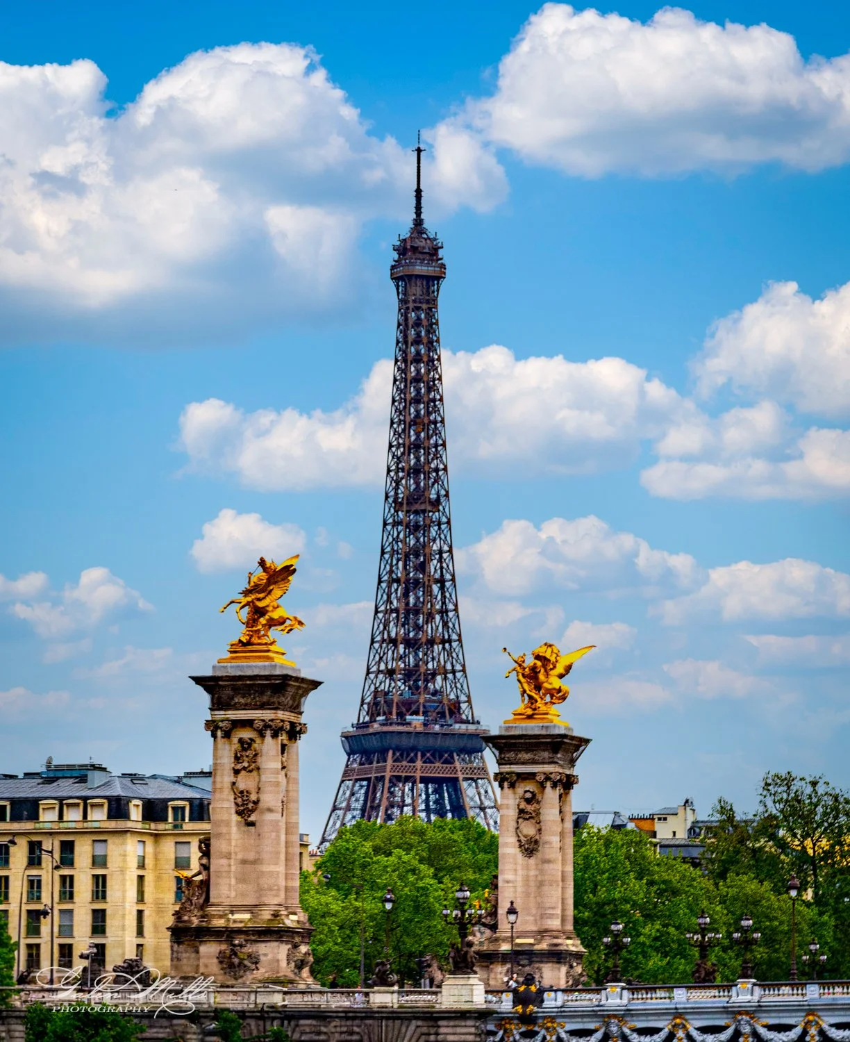 View of the Eiffel Tower with golden statues atop pillars in the foreground, under a blue sky with clouds.