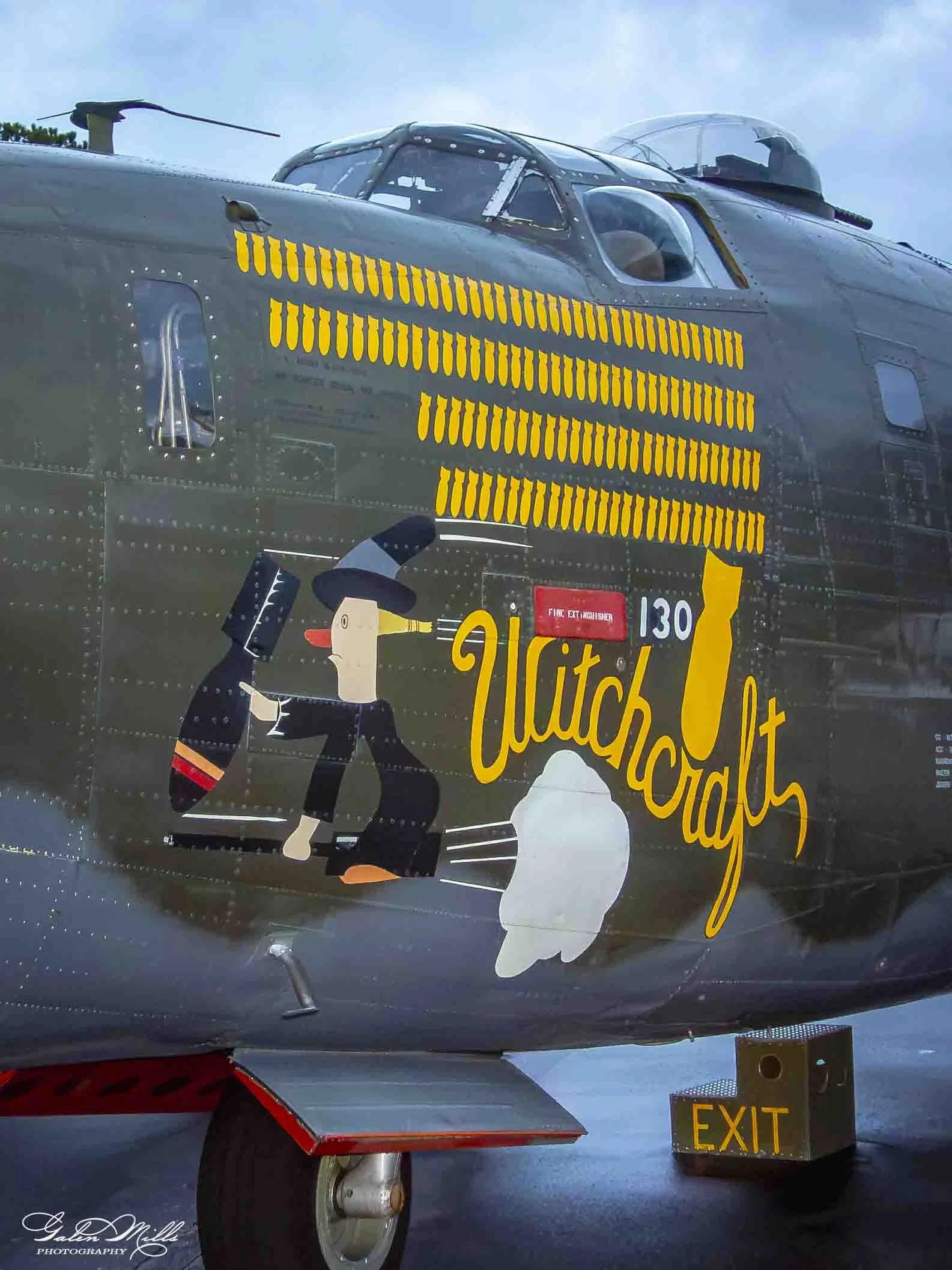 Close-up of a vintage aircraft's nose art featuring a cartoon woman in a black suit with a red headband, holding a large gun, and text that reads 'Dutch' and '130'. The aircraft has a dark gray body with a transparent cockpit canopy and some yellow m