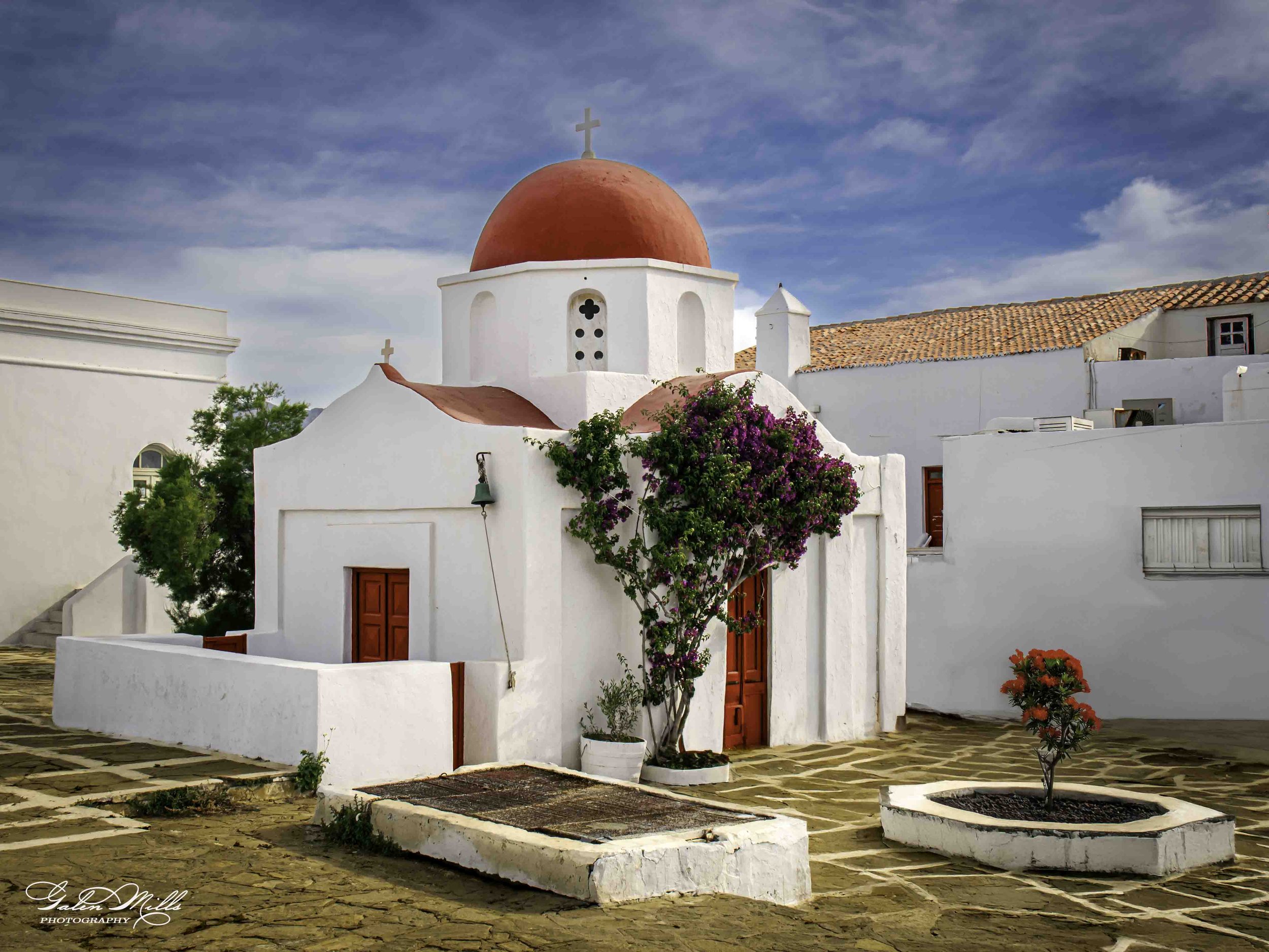 A white chapel with a red domed roof and red doors, surrounded by cobblestone pavement, green plants, and flowering bushes under a blue sky.