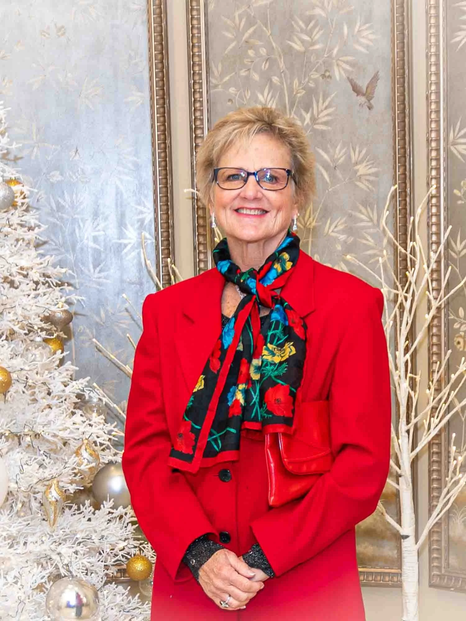 Smiling older woman in a red suit and colorful scarf standing by a white Christmas tree with gold ornaments.