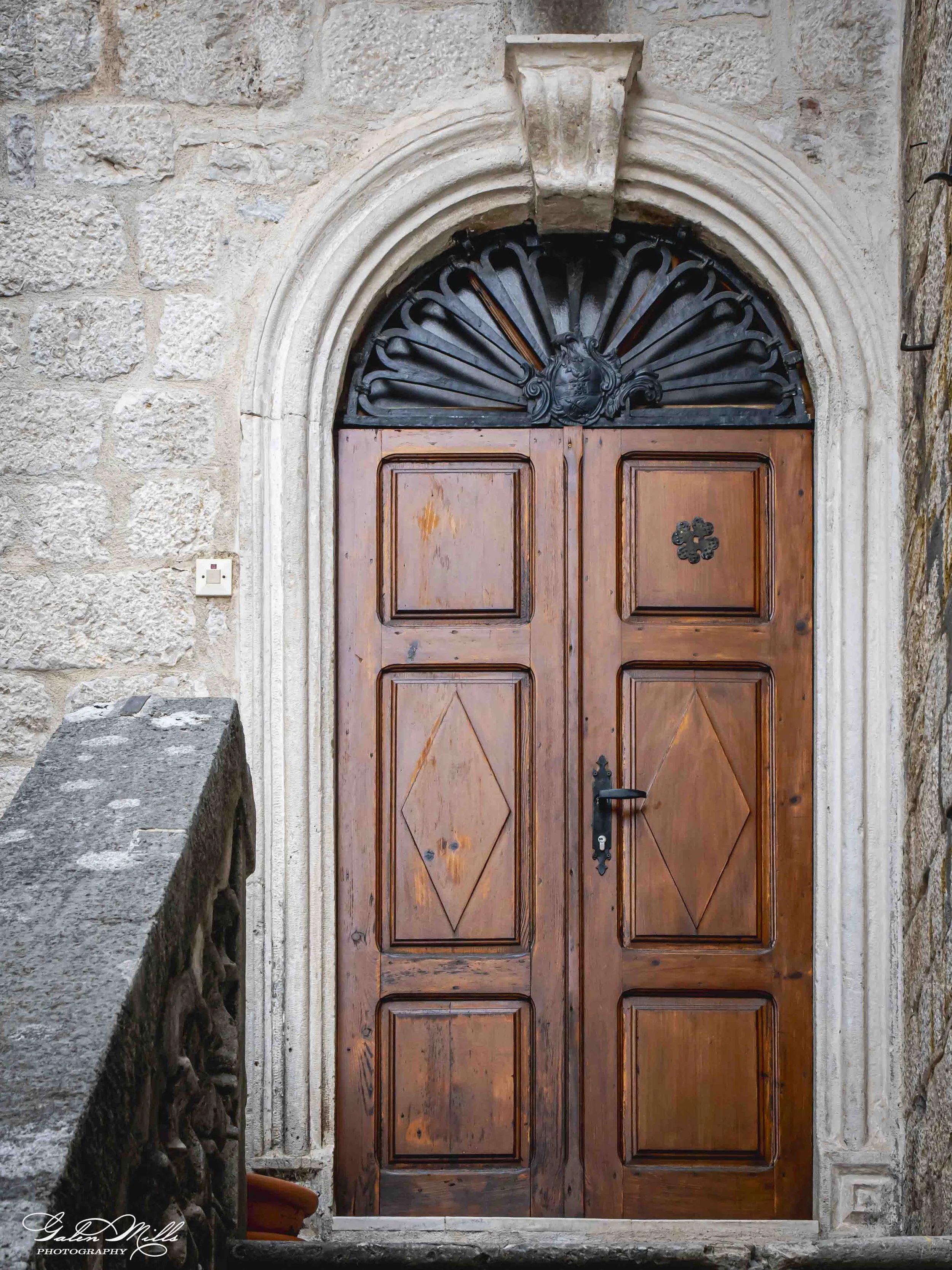 Wooden door with decorative black metalwork and stone arch, set into a stone wall.