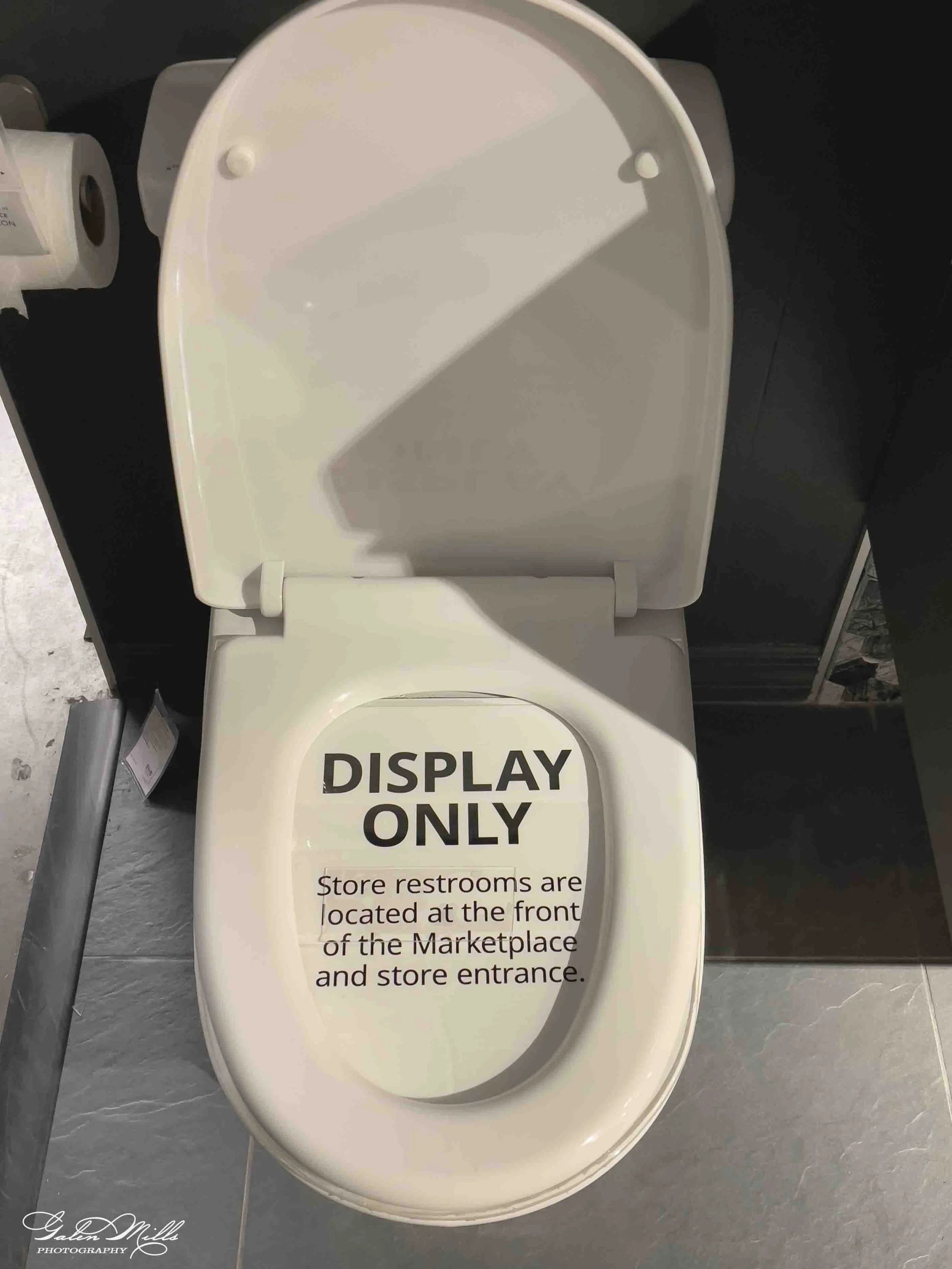 A white ceramic toilet with a sign on the seat that reads, 'DISPLAY ONLY.' A restroom sign and a trash bin are visible nearby.