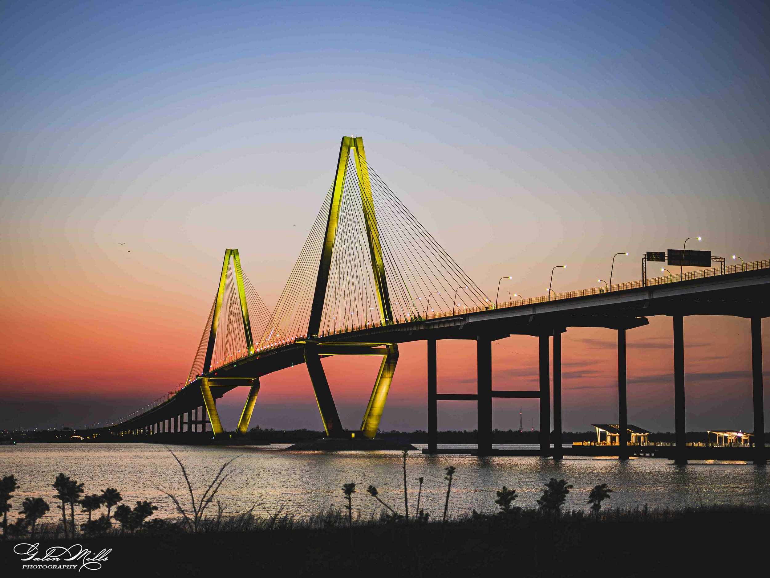 A bridge with illuminated yellow towers and cables spans over water during a sunset or sunrise, with a pastel sky and some trees in the foreground.