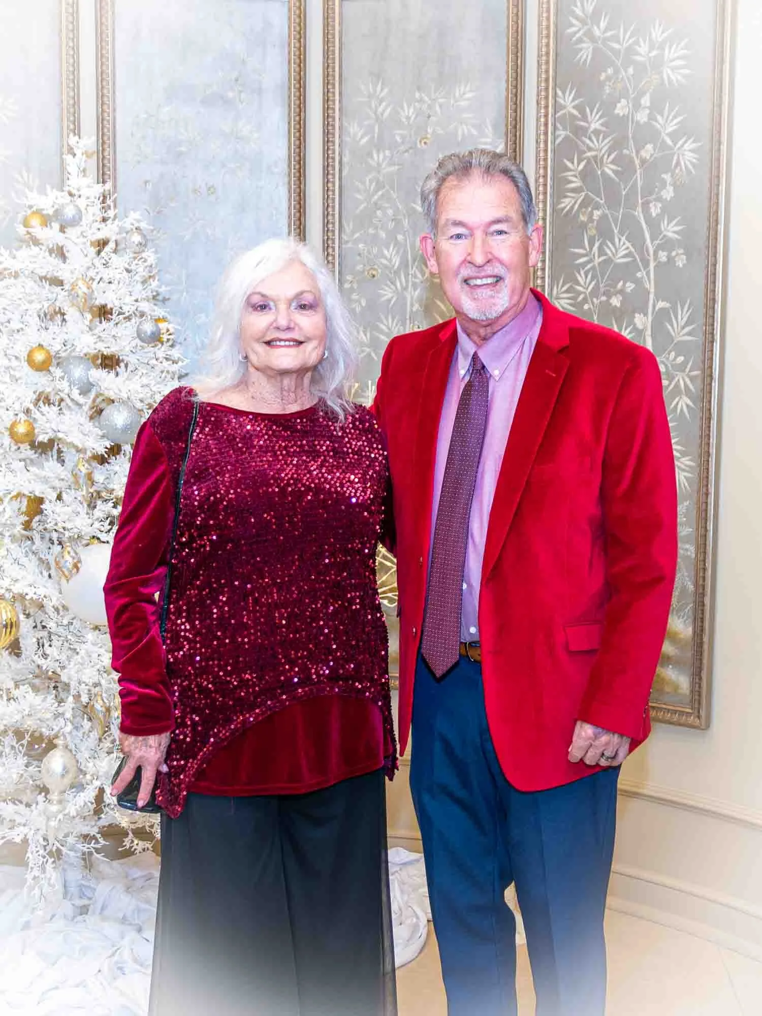 An elderly couple standing in front of a white Christmas tree decorated with gold and silver ornaments. The woman is wearing a red sequined top and black pants, while the man is wearing a red jacket, pink shirt, and tie. The background features elega