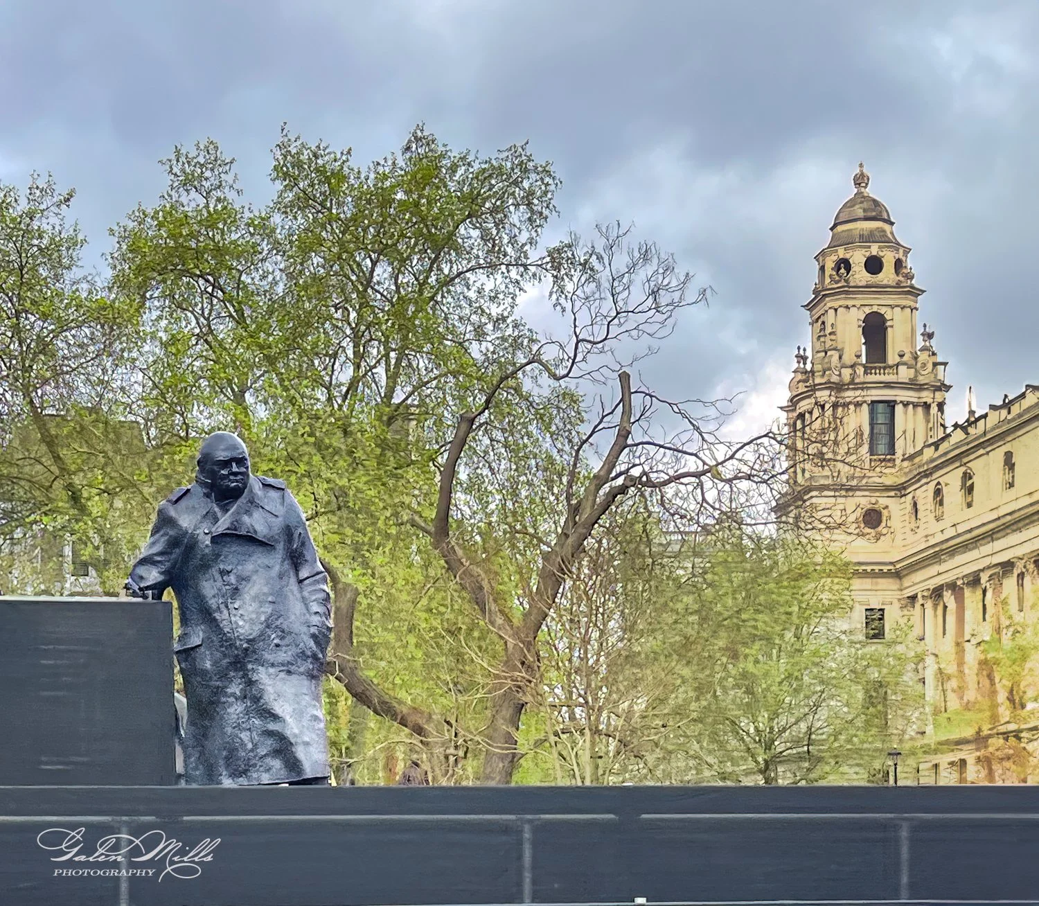 Statue of a figure wearing a coat, surrounded by trees, with a historic building featuring a tall tower in the background, under a cloudy sky.
