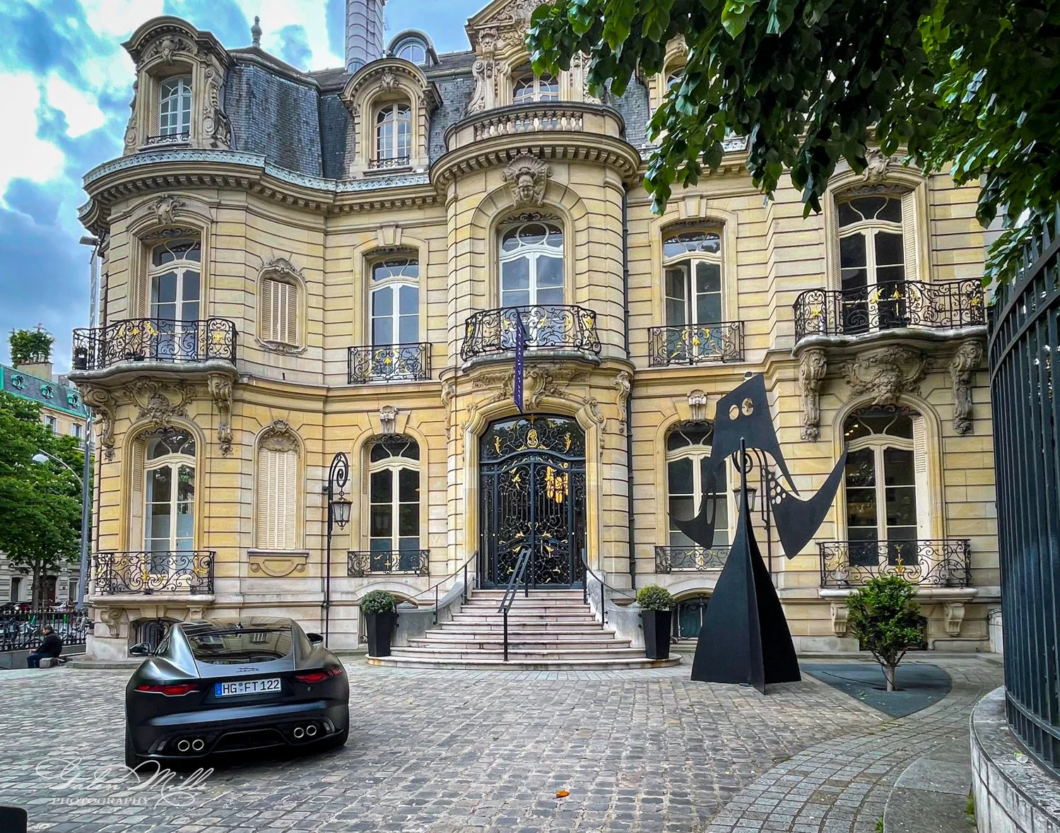 Elegant historic building with ornate architecture featuring wrought iron balconies and large arched windows. A black modern sculpture is displayed in front of the entrance. A luxury car is parked on a stone-paved driveway.