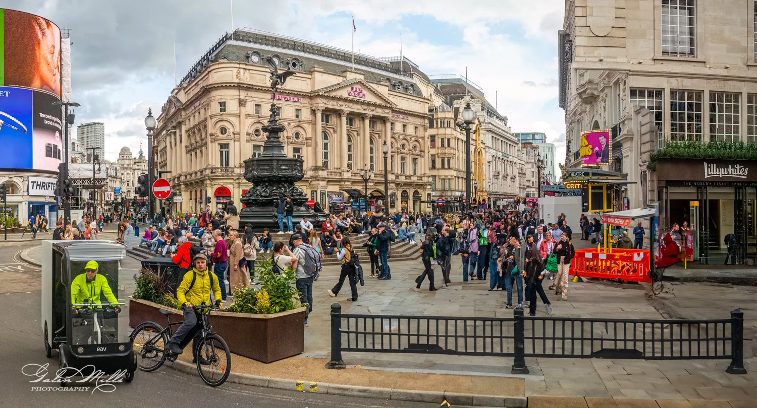Busy urban scene with crowd near historic buildings and a statue in a city square, featuring a cyclist and a mobile delivery cart.