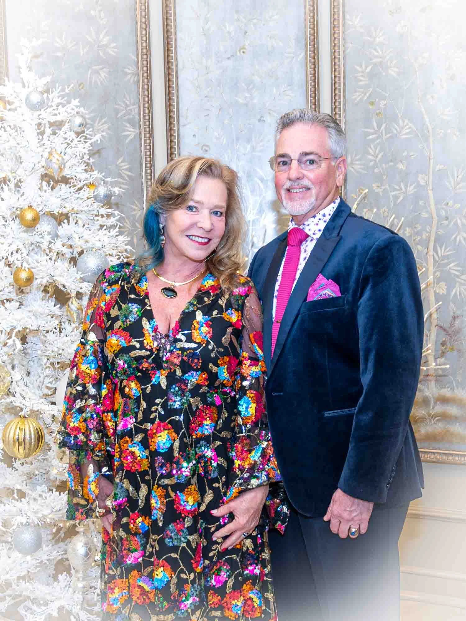 A woman in a colorful floral dress and a man in a suit with a pink tie stand in front of a white Christmas tree with gold and silver ornaments.