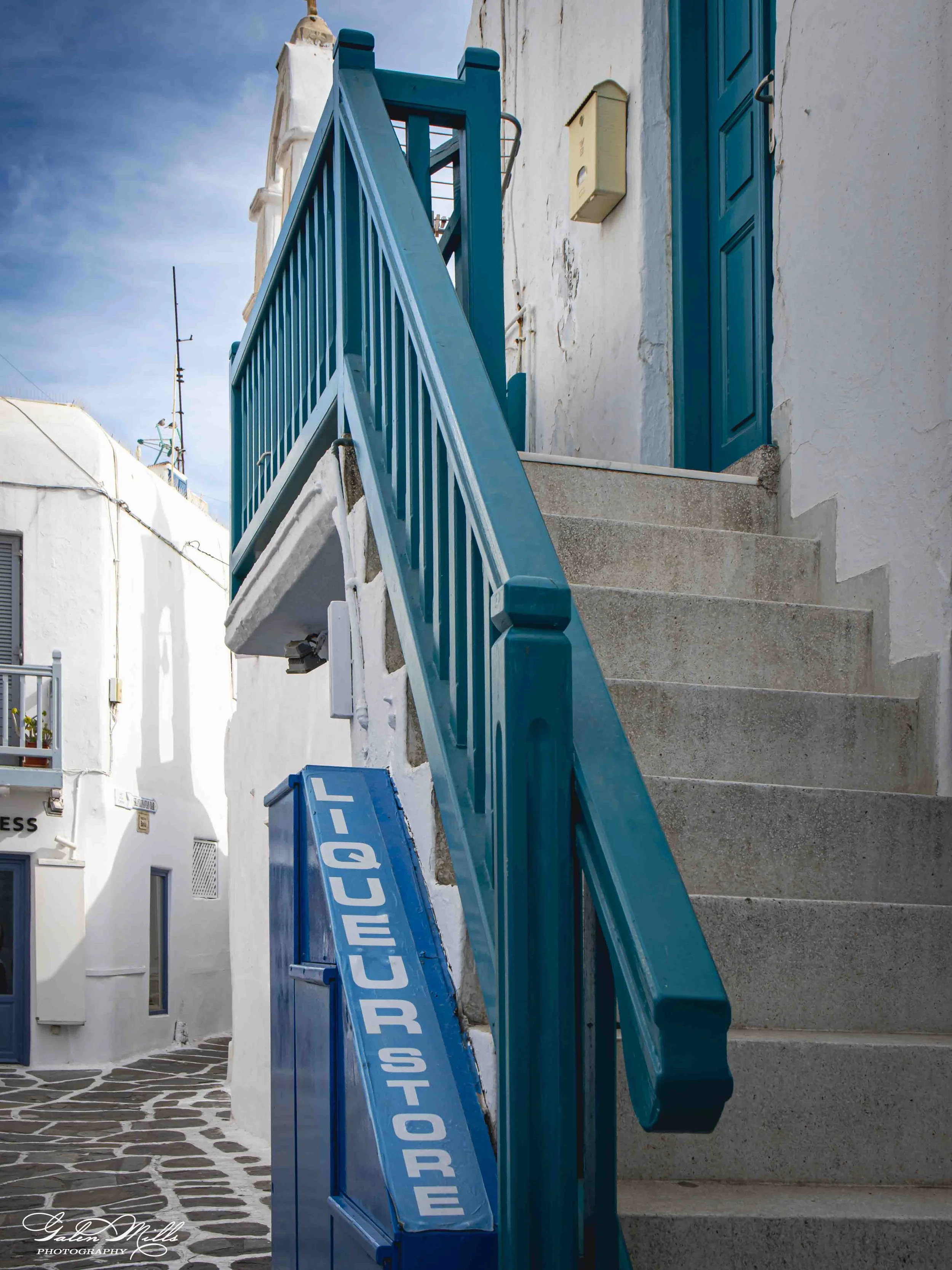 Blue sign with white letters that reads 'QUIET PLEASE', mounted on a wall beside a staircase leading to a blue door, with a white building in the background and clear sky above.