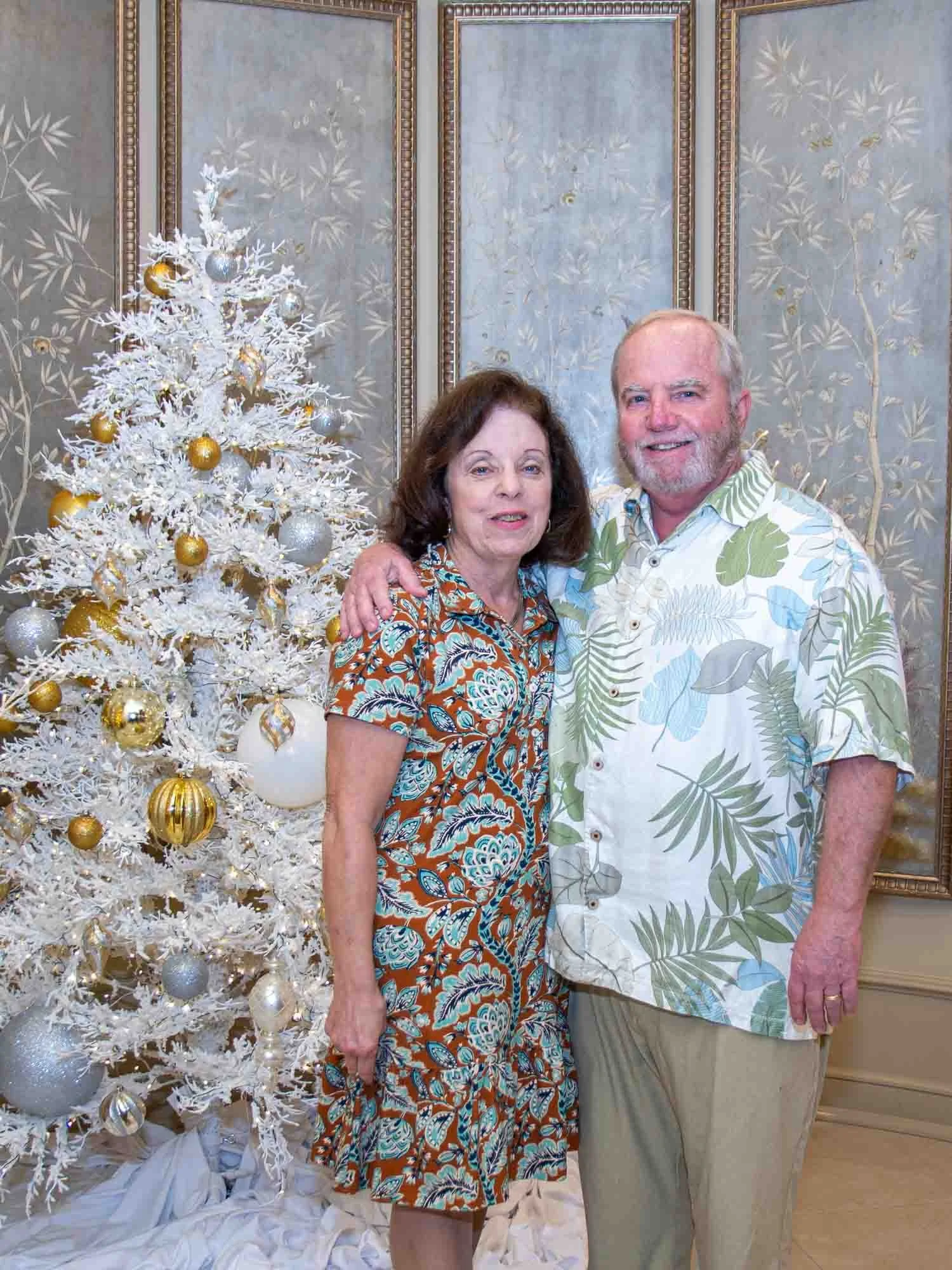 A couple standing in front of a white Christmas tree decorated with gold and silver ornaments. The woman is wearing a floral dress, and the man is in a tropical shirt. Ornate wallpaper serves as the background.