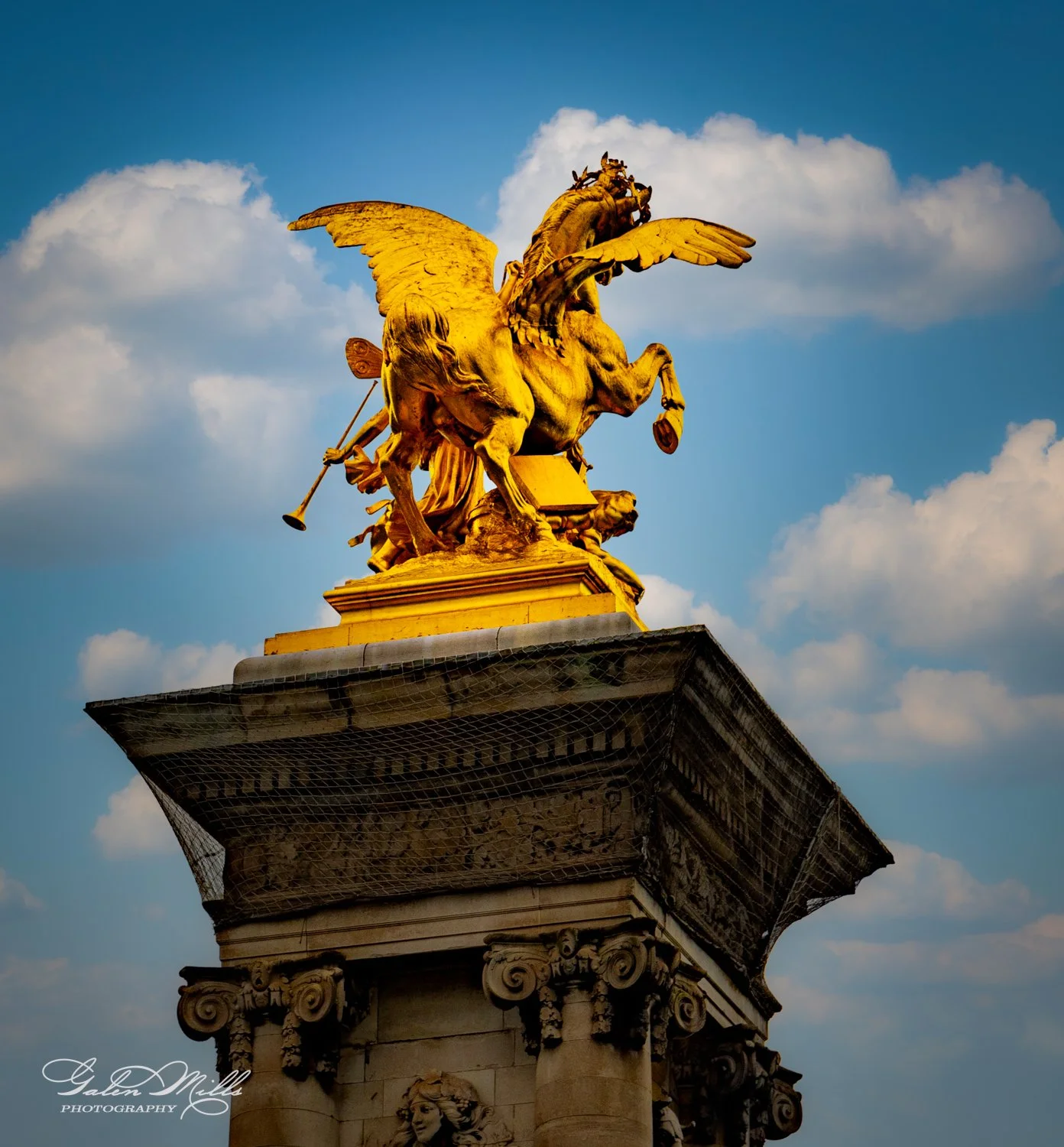 Golden statue of a winged horse with a human figure, possibly on a bridge, against a blue sky with clouds.