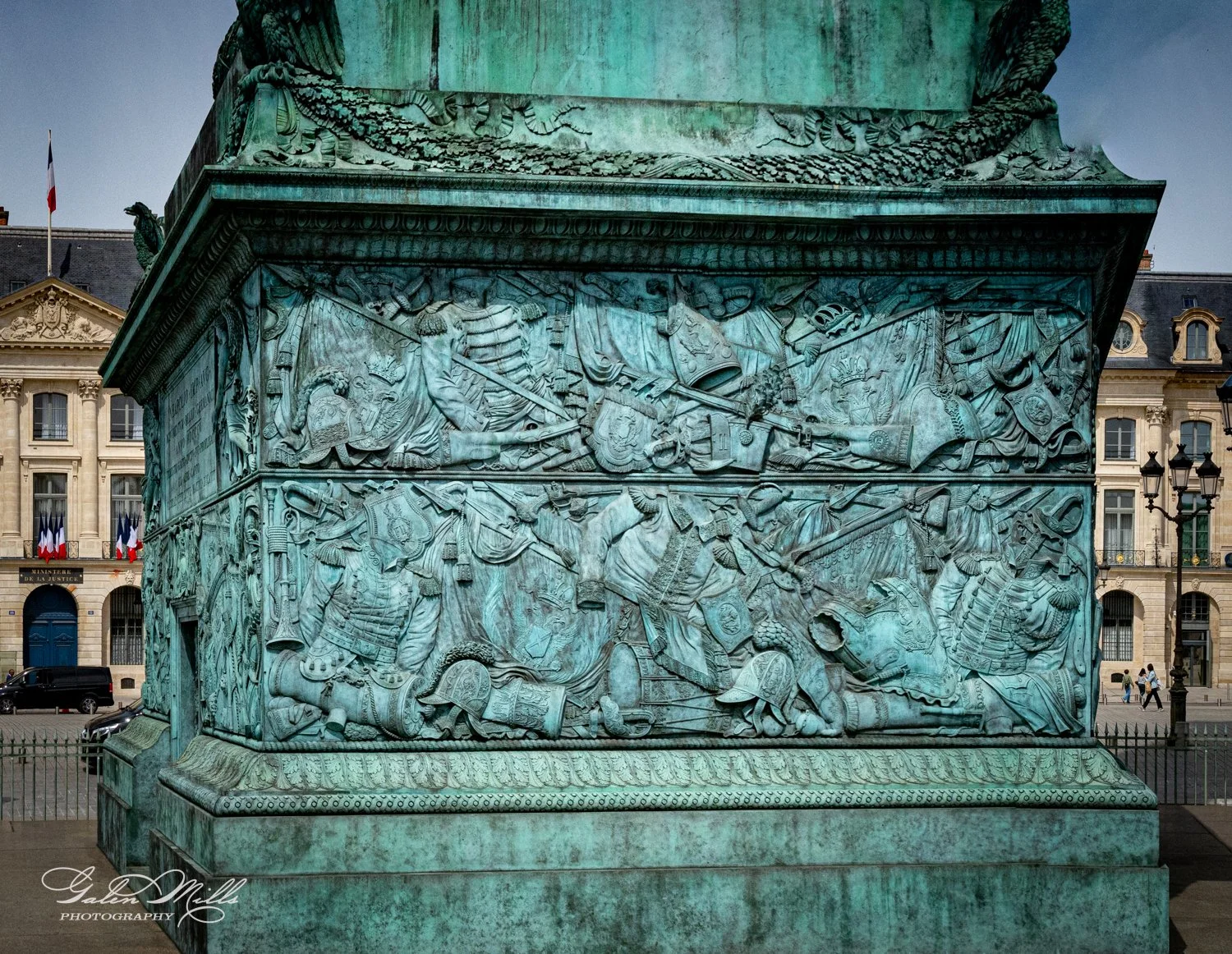 Close-up of bronze relief depicting battle scenes, part of a monument in a city square, with historic buildings and French flags in the background.
