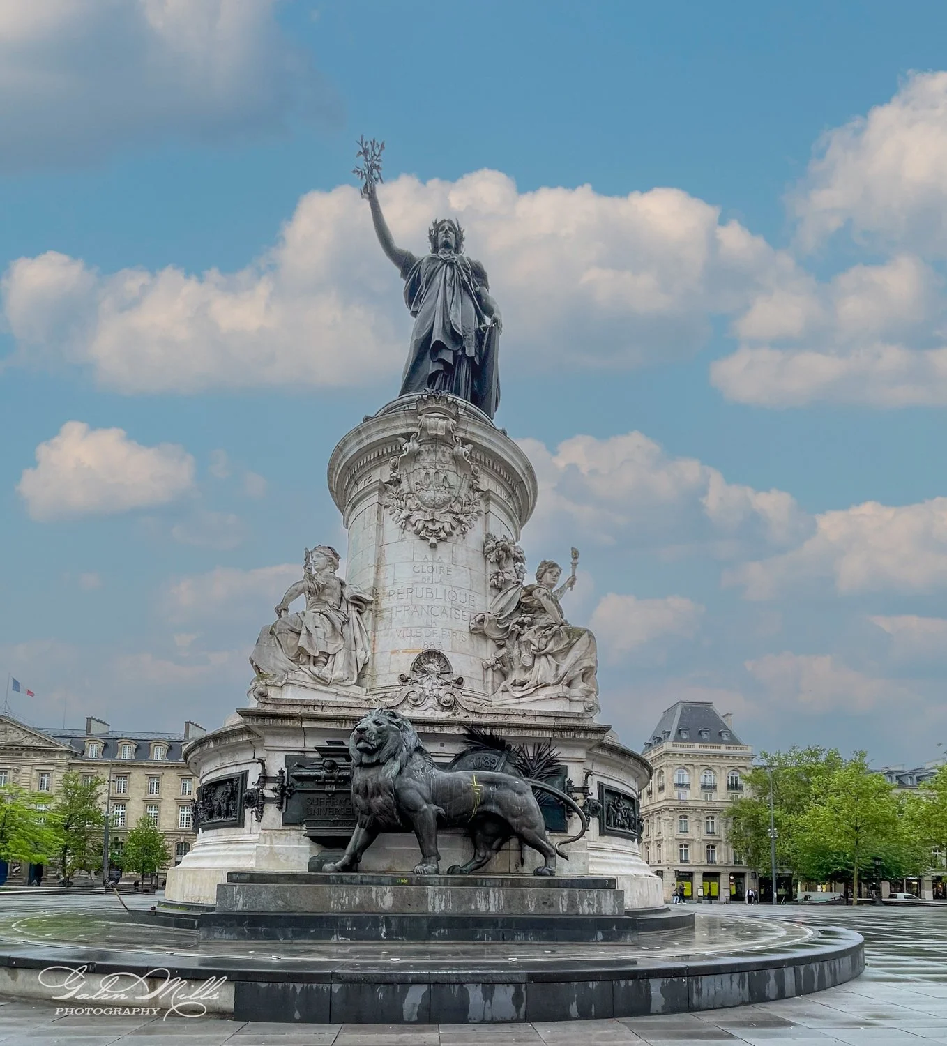 Statue at Place de la République, Paris, featuring Marianne holding a branch, flanked by sculptures and a lion on a pedestal in a public square.