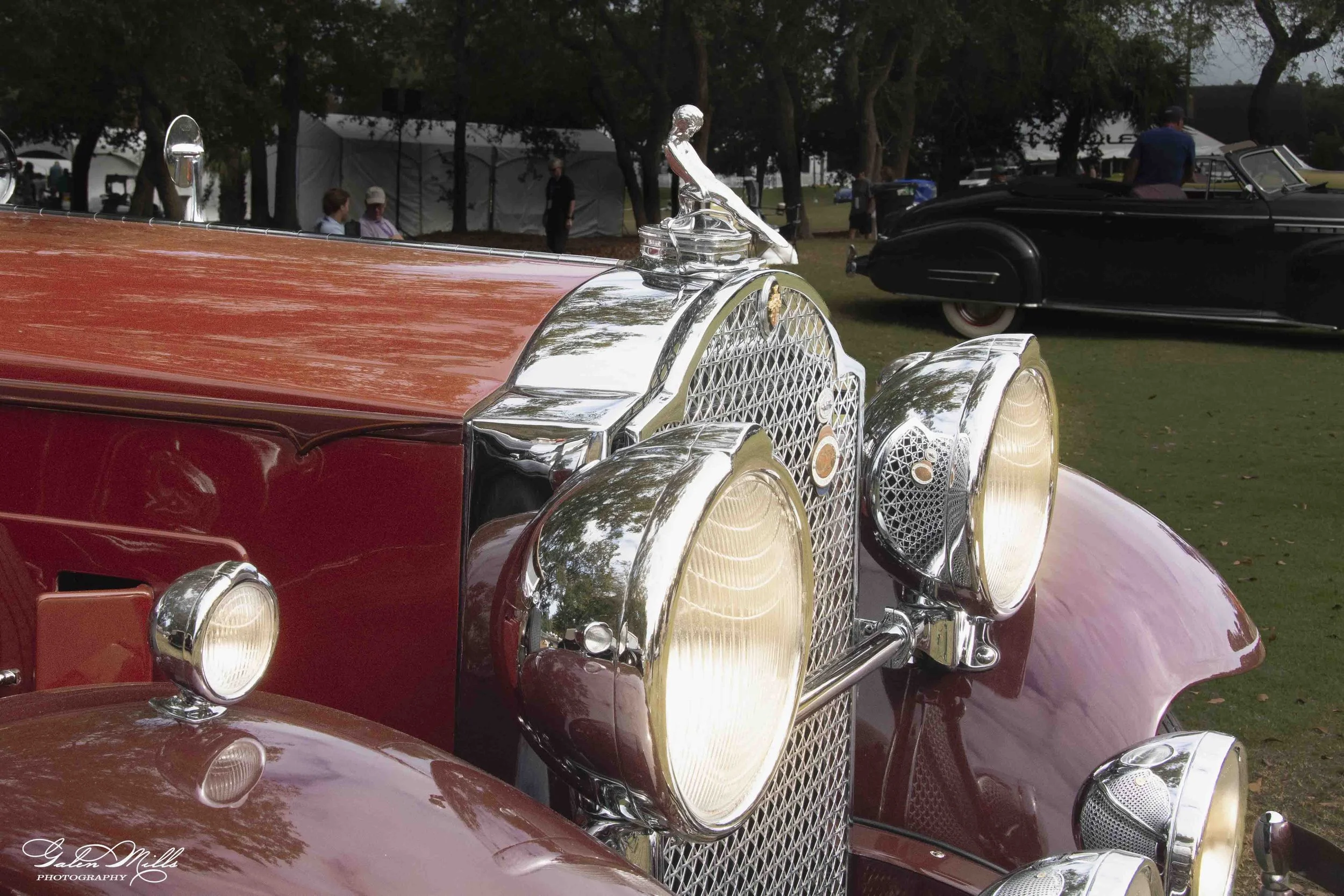 Close-up of a vintage red car with shiny chrome details, including a hood ornament and front grille, at an outdoor classic car exhibition.