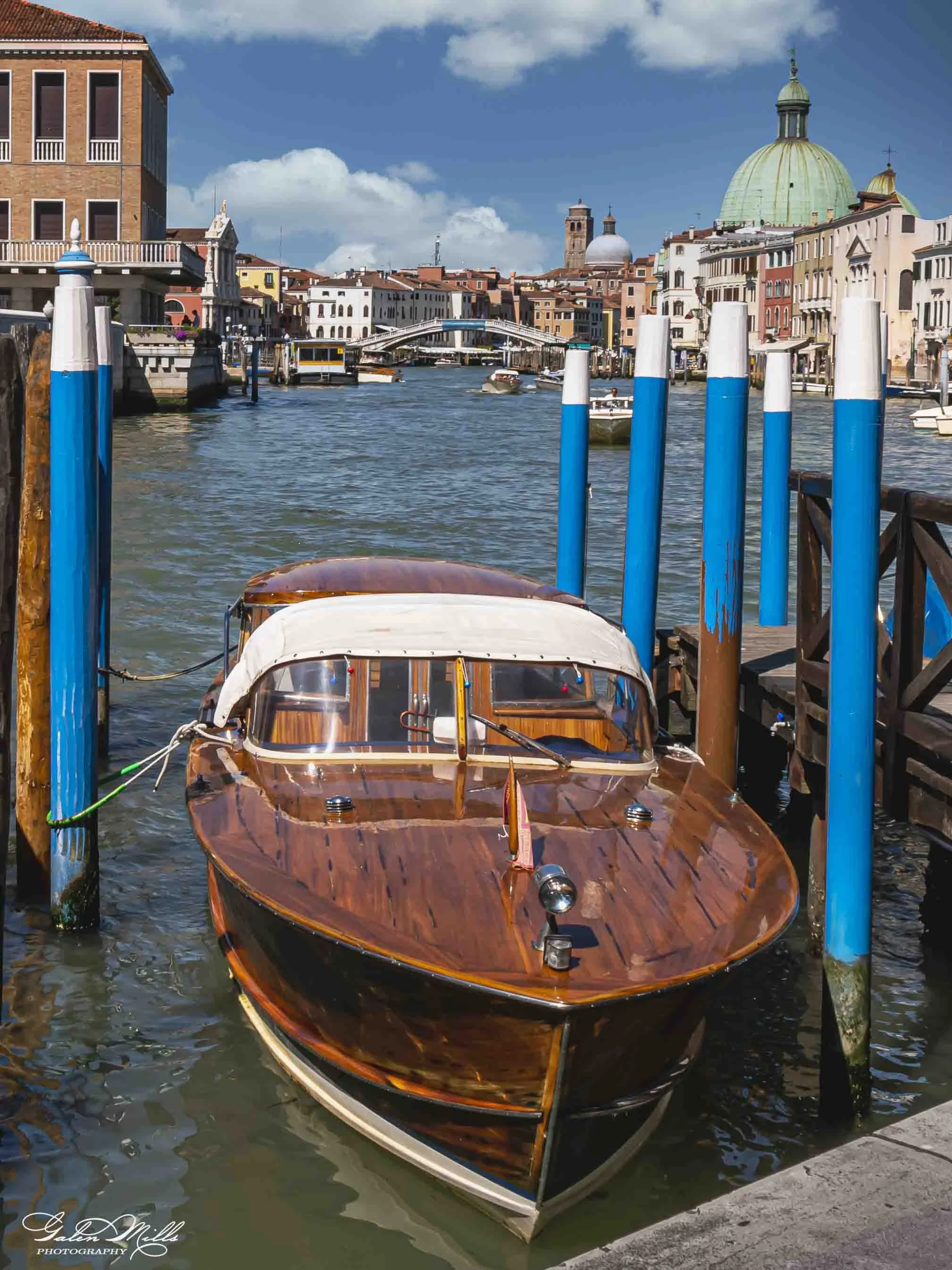 A wooden motorboat docked at a small pier in a canal with blue and white striped posts in Venice, Italy, with colorful historic buildings and domed churches in the background.