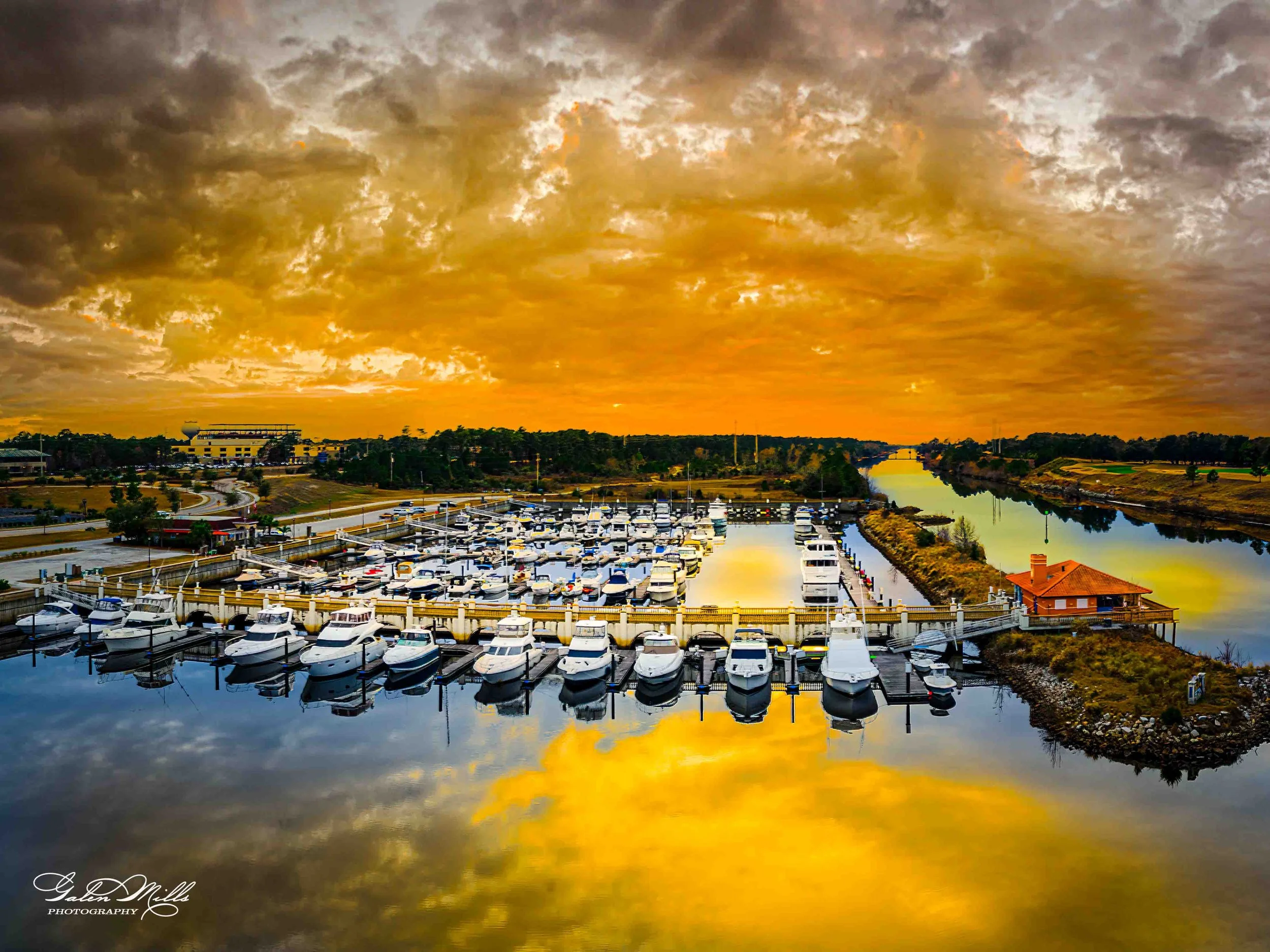 A marina filled with boats at sunset, with a dramatic orange and yellow sky reflected in the water, and a small building on a pier on the right side.