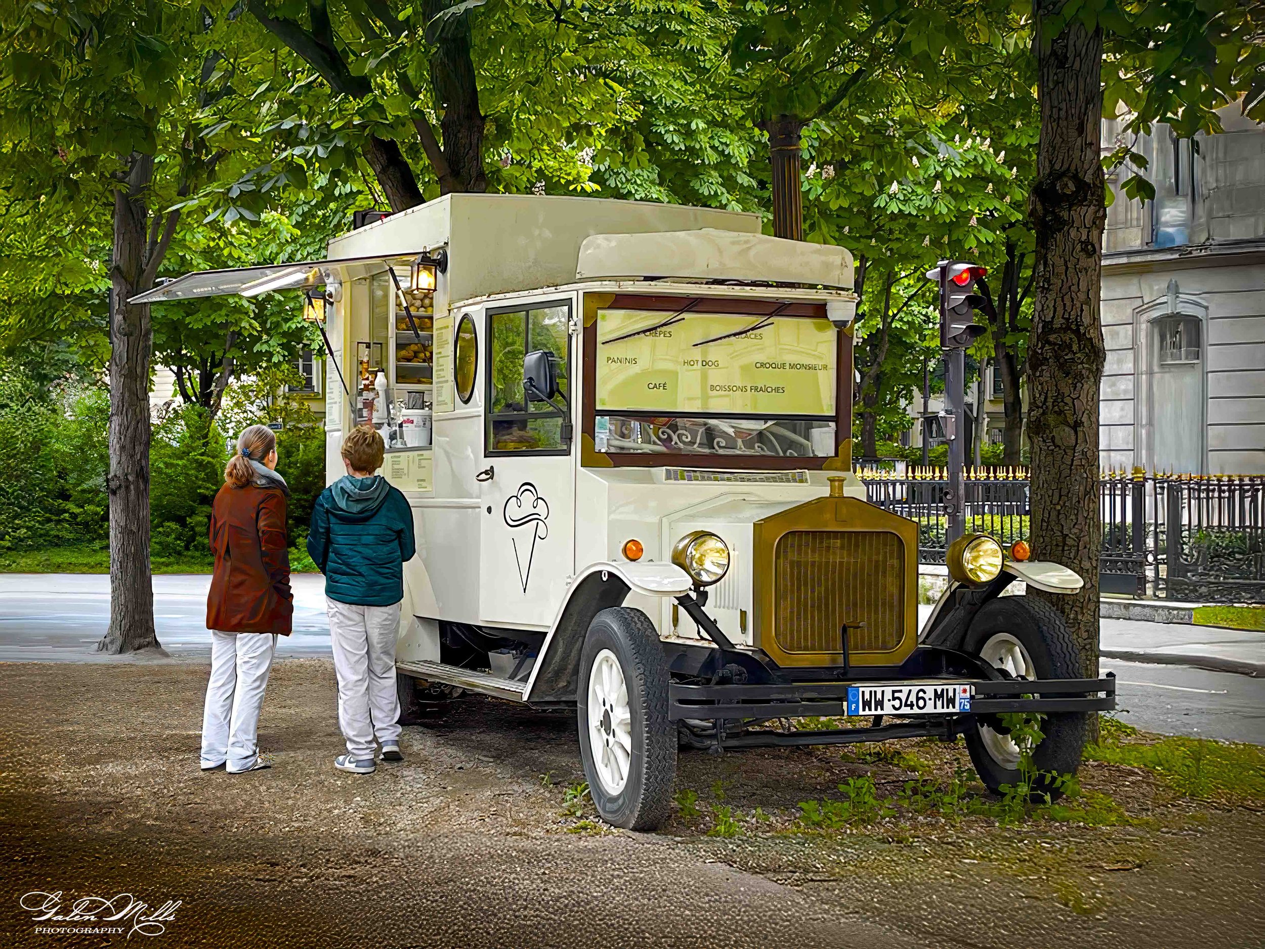 Vintage ice cream truck with two people, one wearing a coat, parked beneath leafy trees on a street. The truck has a menu and ice cream cone illustration on the side.
