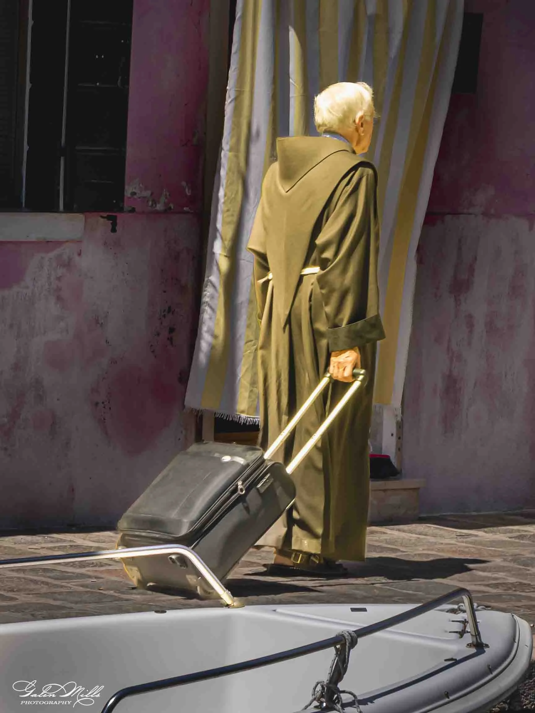 An elderly person with white hair, wearing a tan coat, stands next to a large window with striped curtains, holding a cane, with a black rolling suitcase placed on the ground beside them. In the foreground, there is a white inflatable boat with black