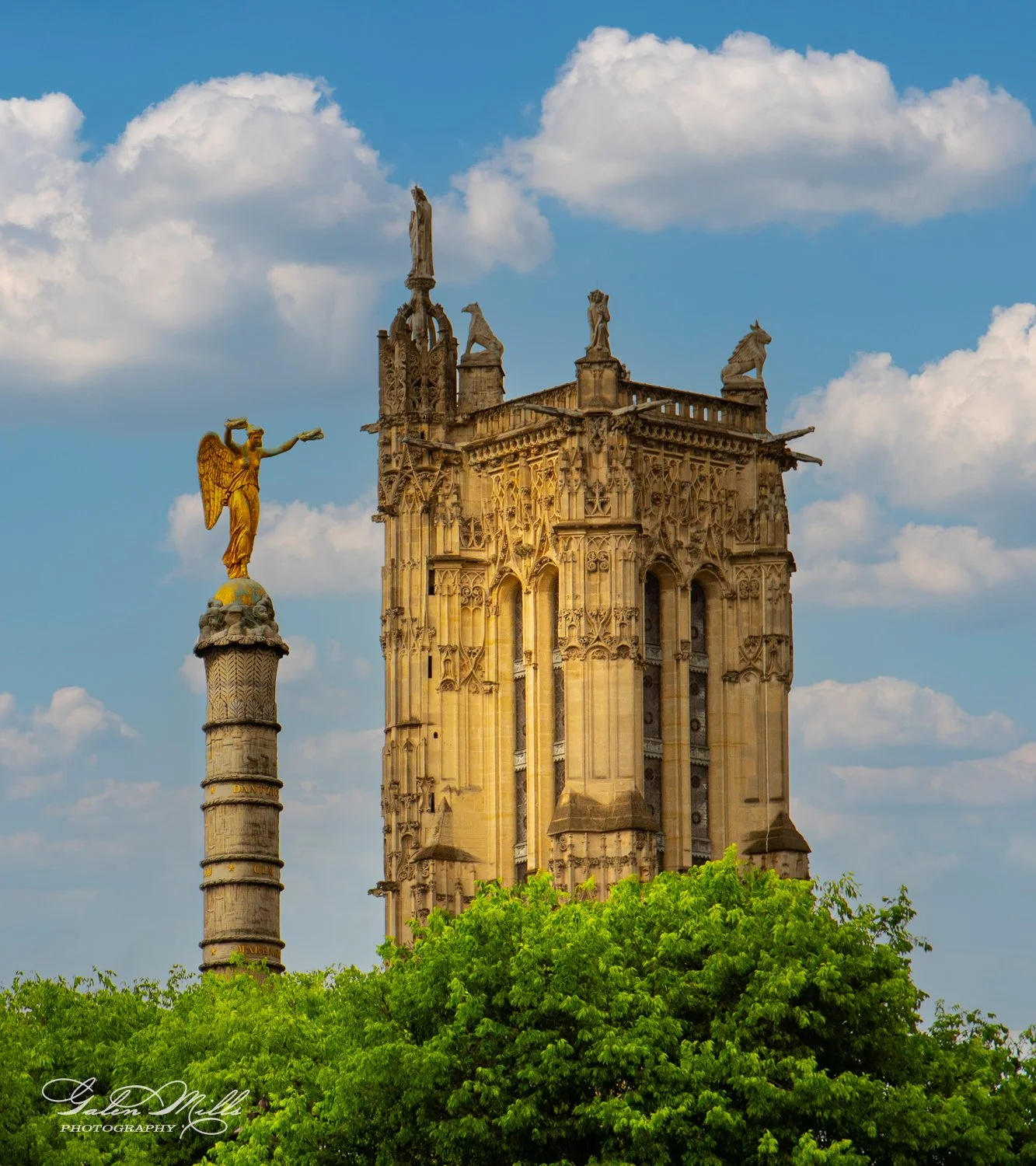 Historic stone tower and golden angel statue under a blue sky with clouds, surrounded by green trees.