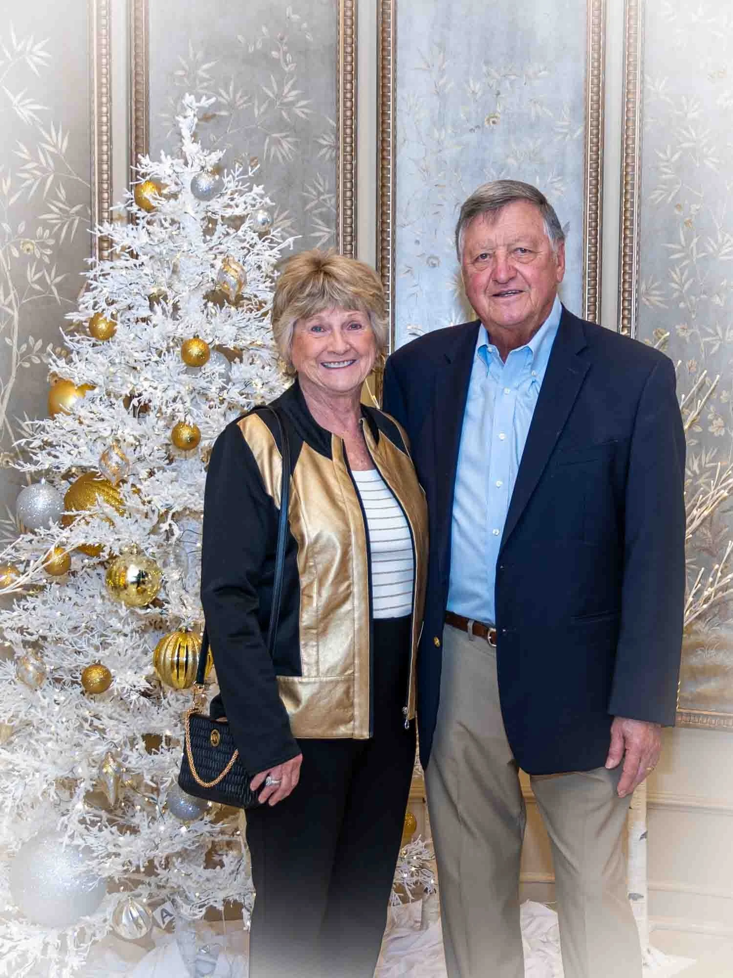 An older couple smiling in front of a white Christmas tree decorated with gold and silver ornaments. The woman is wearing a gold and black jacket, and the man is in a dark blazer and light blue shirt.