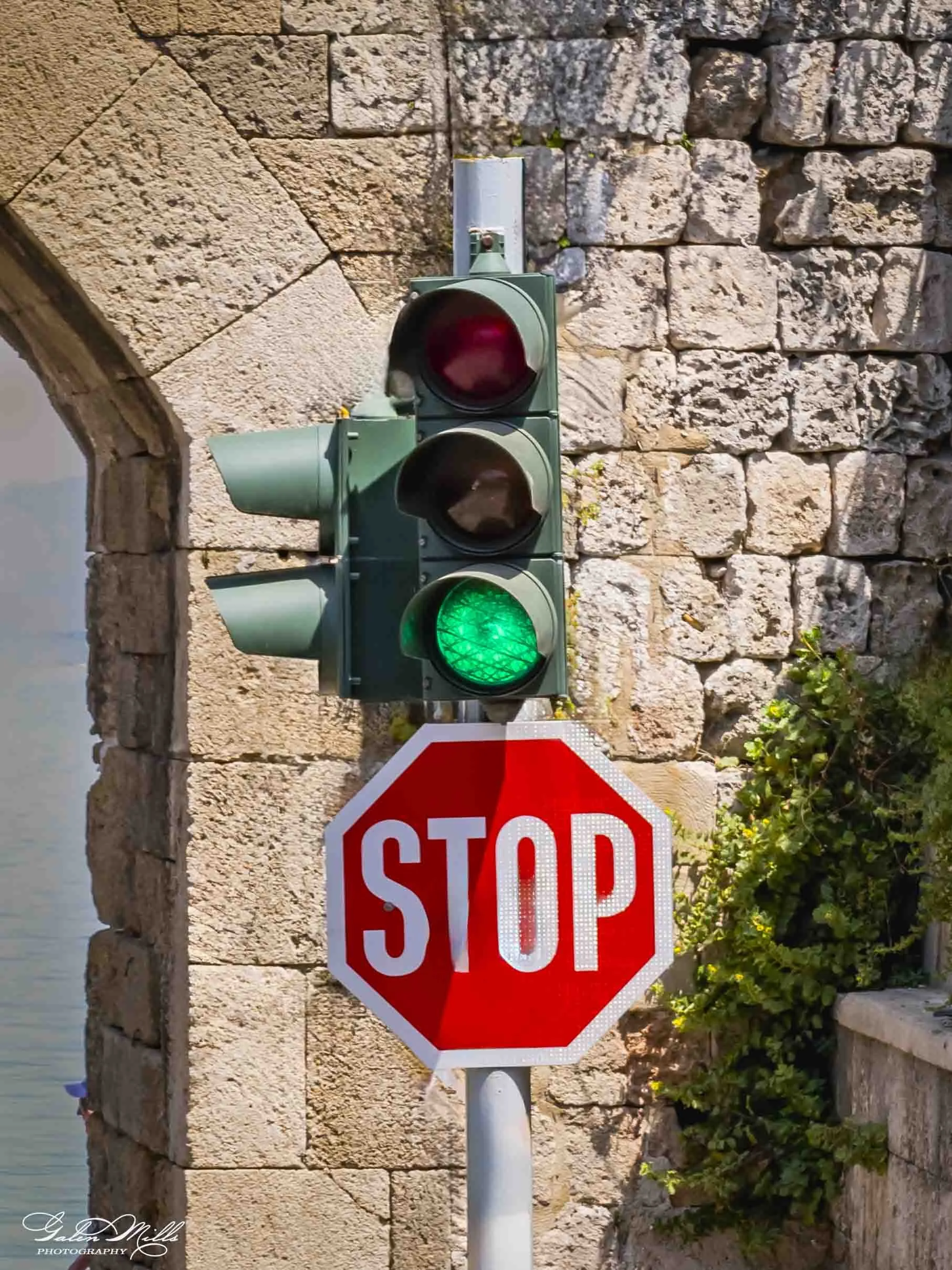 A traffic light displaying a green signal and a stop sign attached underneath, in front of an old stone wall with some greenery.