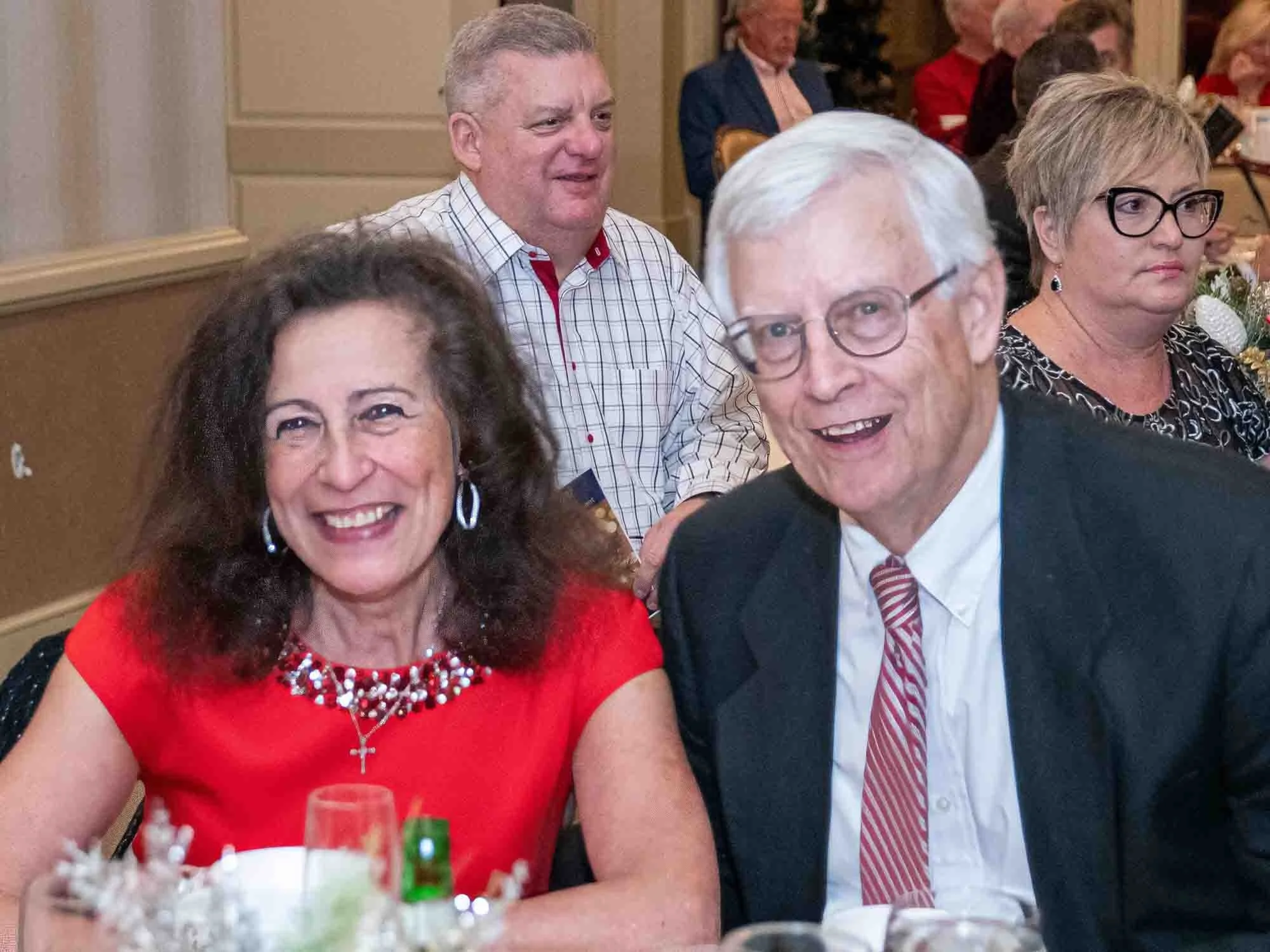A group of people sitting at a formal event, with a woman in a red dress and a man in a suit smiling at the camera.