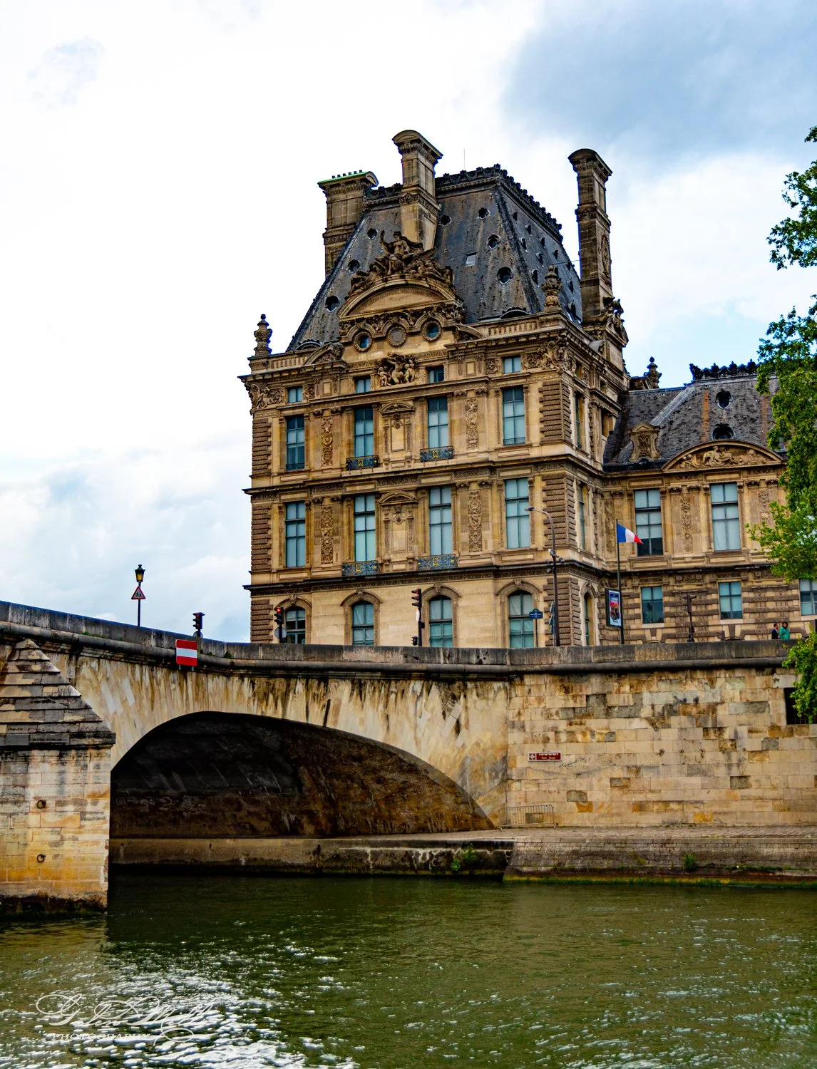 Historic building with ornate architecture next to a stone bridge over a river, under a cloudy sky.