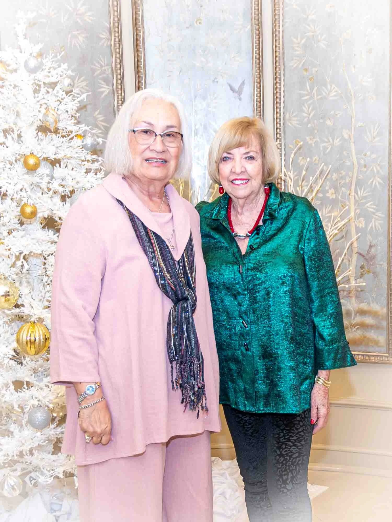 Two women standing indoors, one in a pink outfit with a scarf and the other in a green blouse, near a white Christmas tree decorated with gold ornaments.