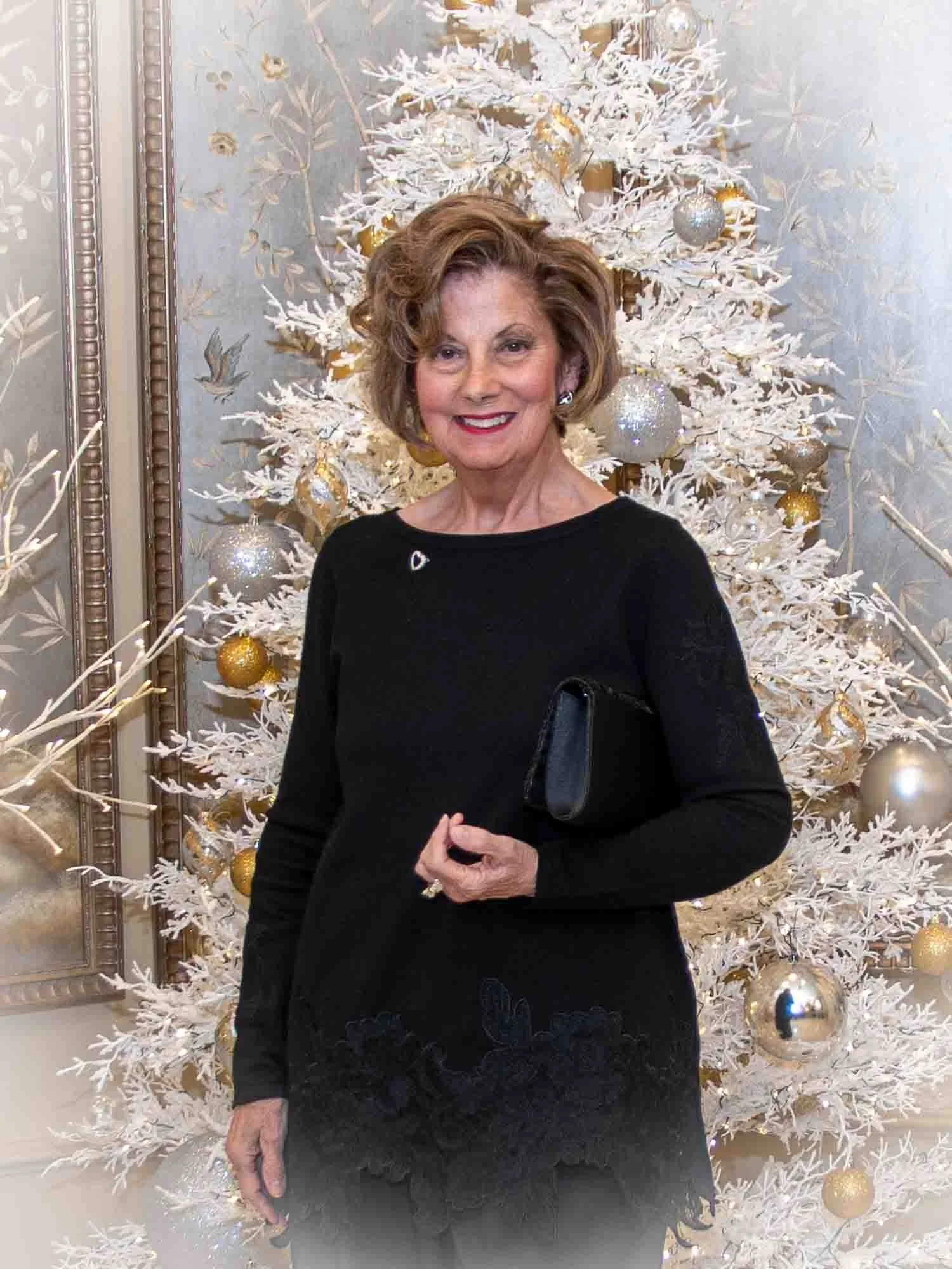 A woman stands in front of a white Christmas tree decorated with gold and silver ornaments, wearing a black outfit and holding a black clutch.