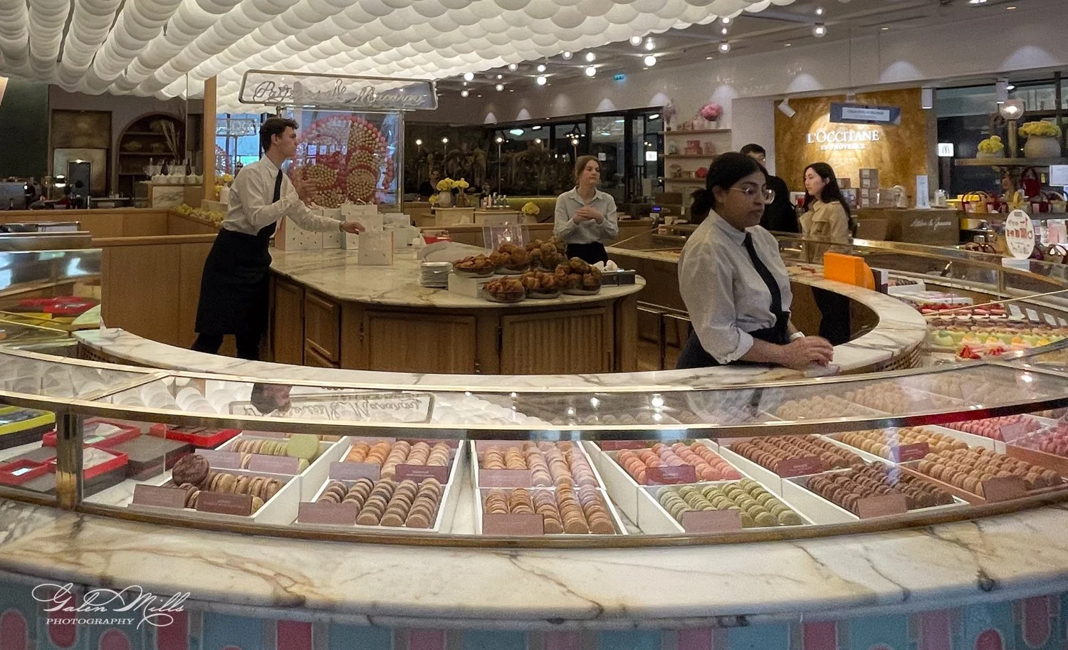Interior of a pastry shop with a counter display of colorful macarons. Staff are attending to customers amid a bright, elegant decor.